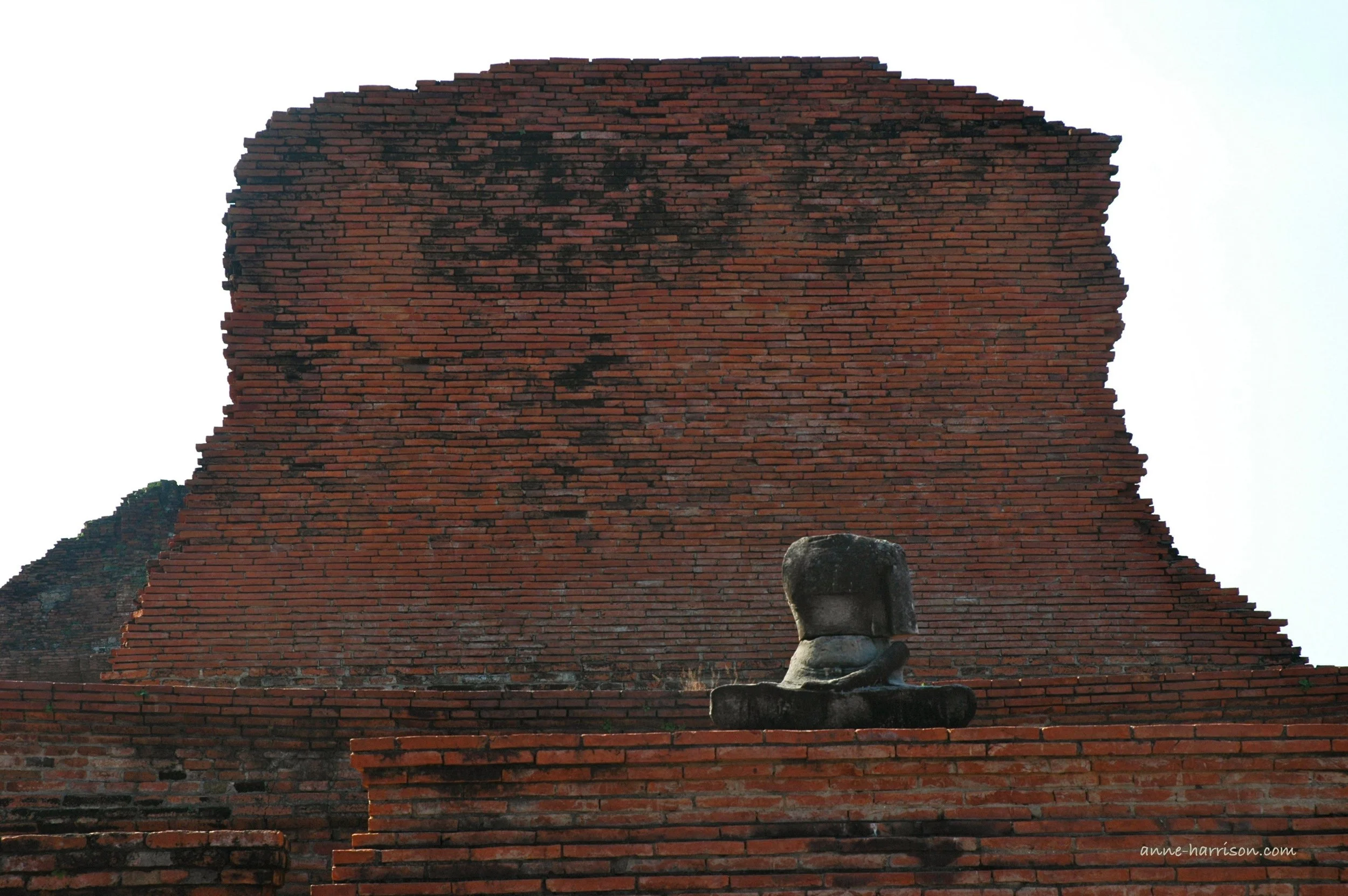 A ruined statue of a buddha, with only one arm and no head, sits infront of a ruined brick wall which was once part of a temple