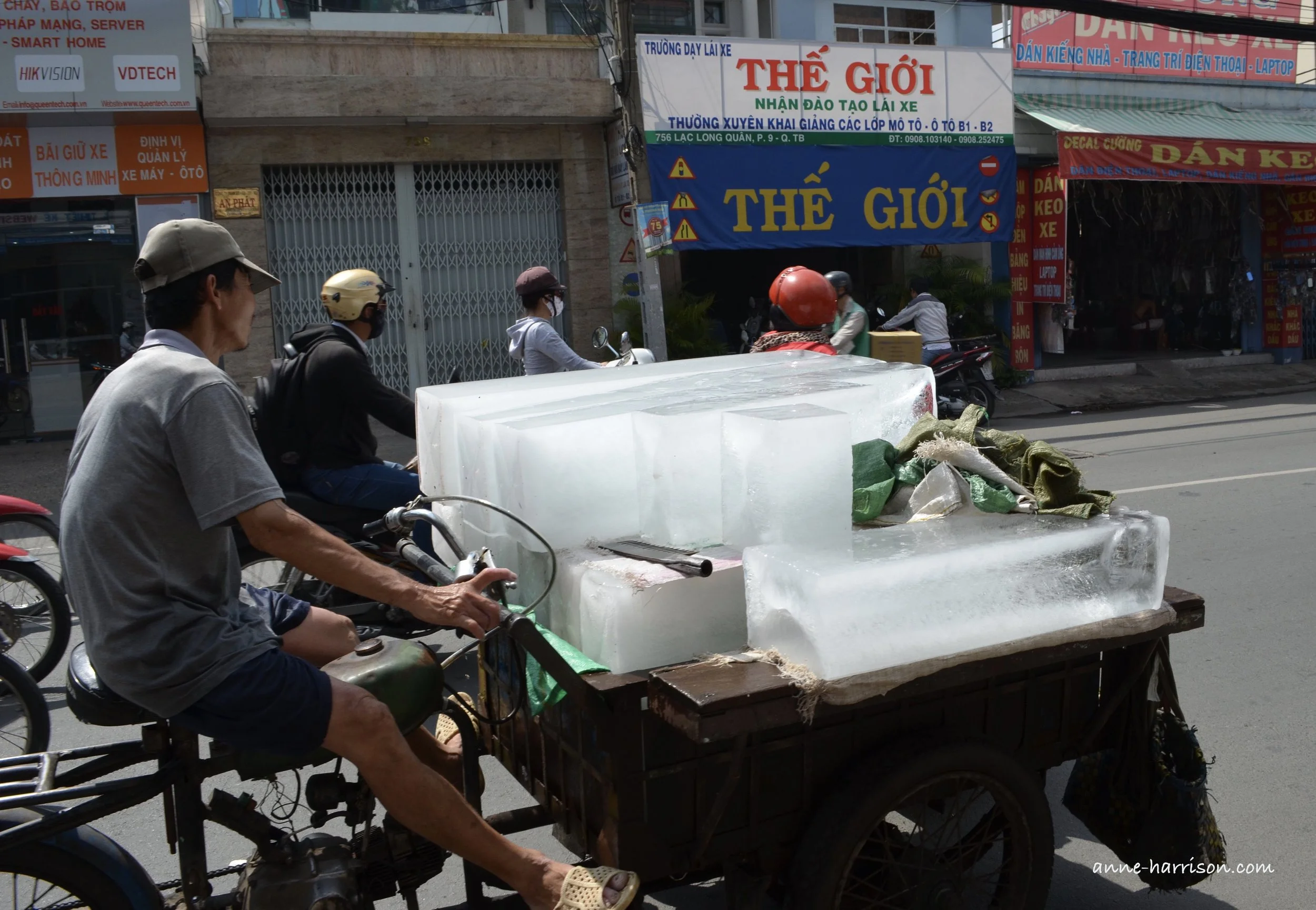 A man on a motor bike with a trailer over the front wheel, carrying blocks of ice. The street is lines with shops and signs in Vietnamese.