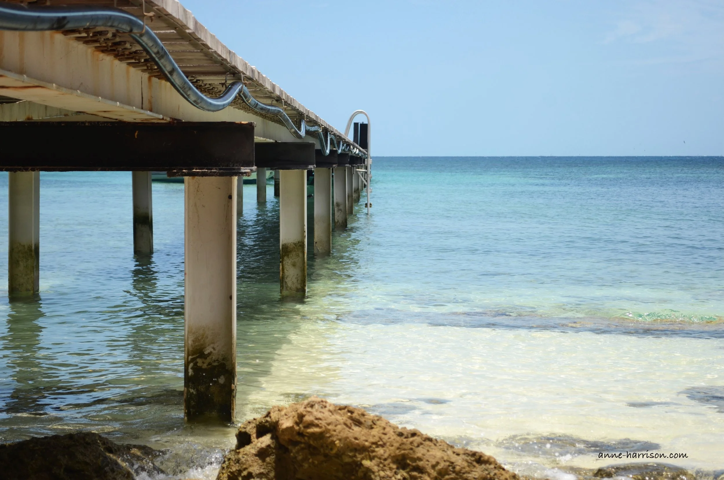 A wooden jetty stretching into tranquil blue water. The sky is also blue.