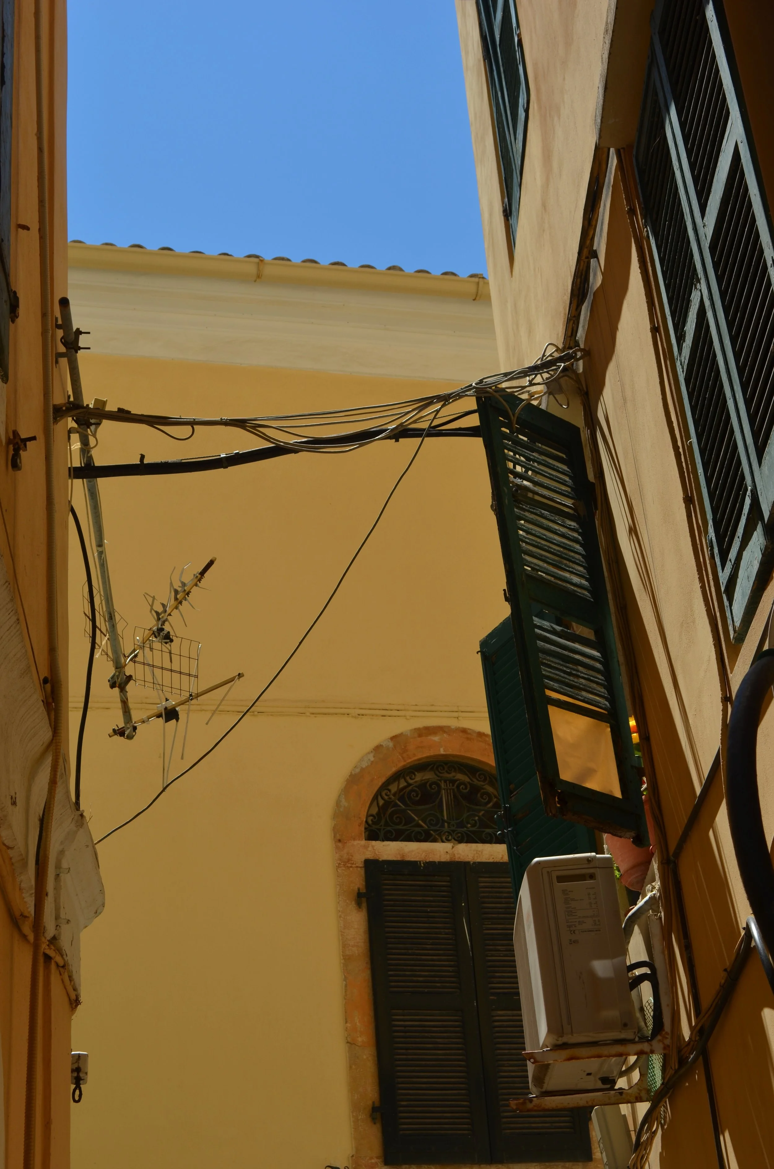  Details of apartment buildings in Corfu, Greece. They are yellow, with black shutters. One window shutter is open. Telephone and electricity wires connect the buildings, and one has an antenna. 