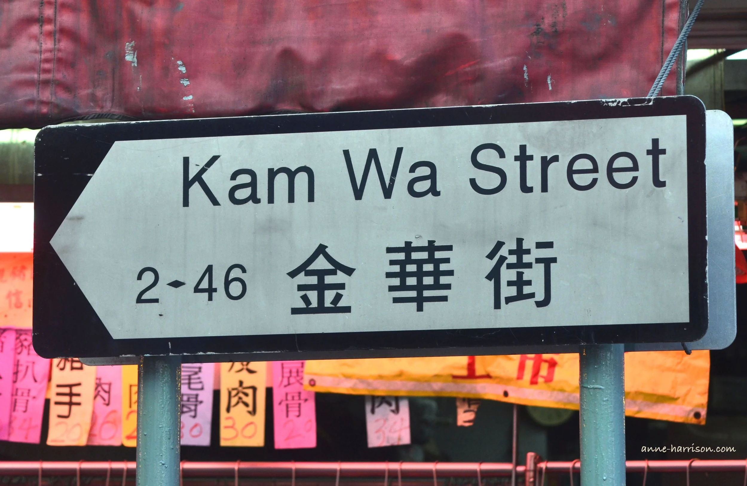 A road sign in Hong Kong saying Kam Wa Street in English and in Cantonese. Colourful signs are behind it.