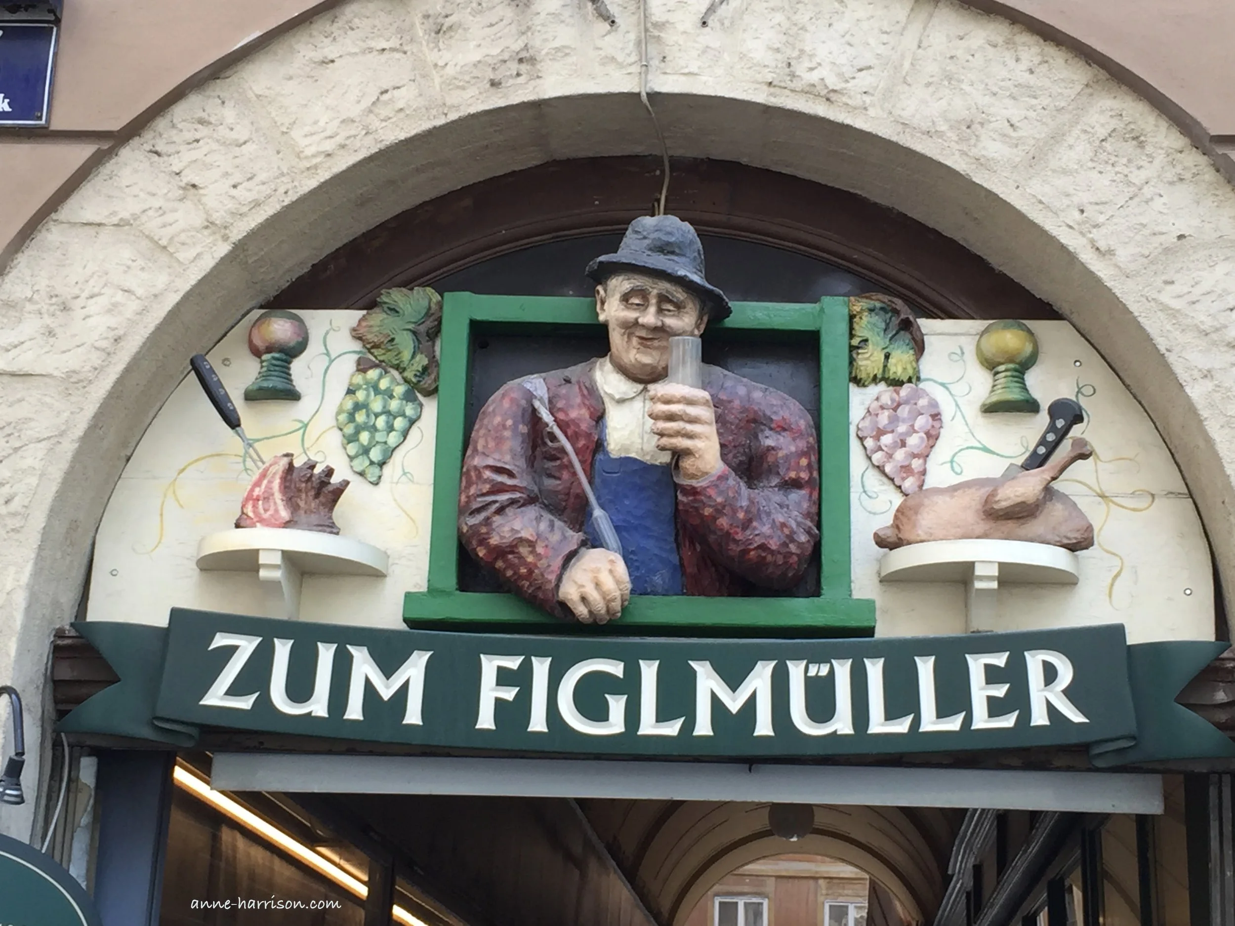 A colourful sign above the entrance to a beer hall in Vienna. A aan in a hat holds a glass, and beside him are images of grapes, and meat ready to be fooked.