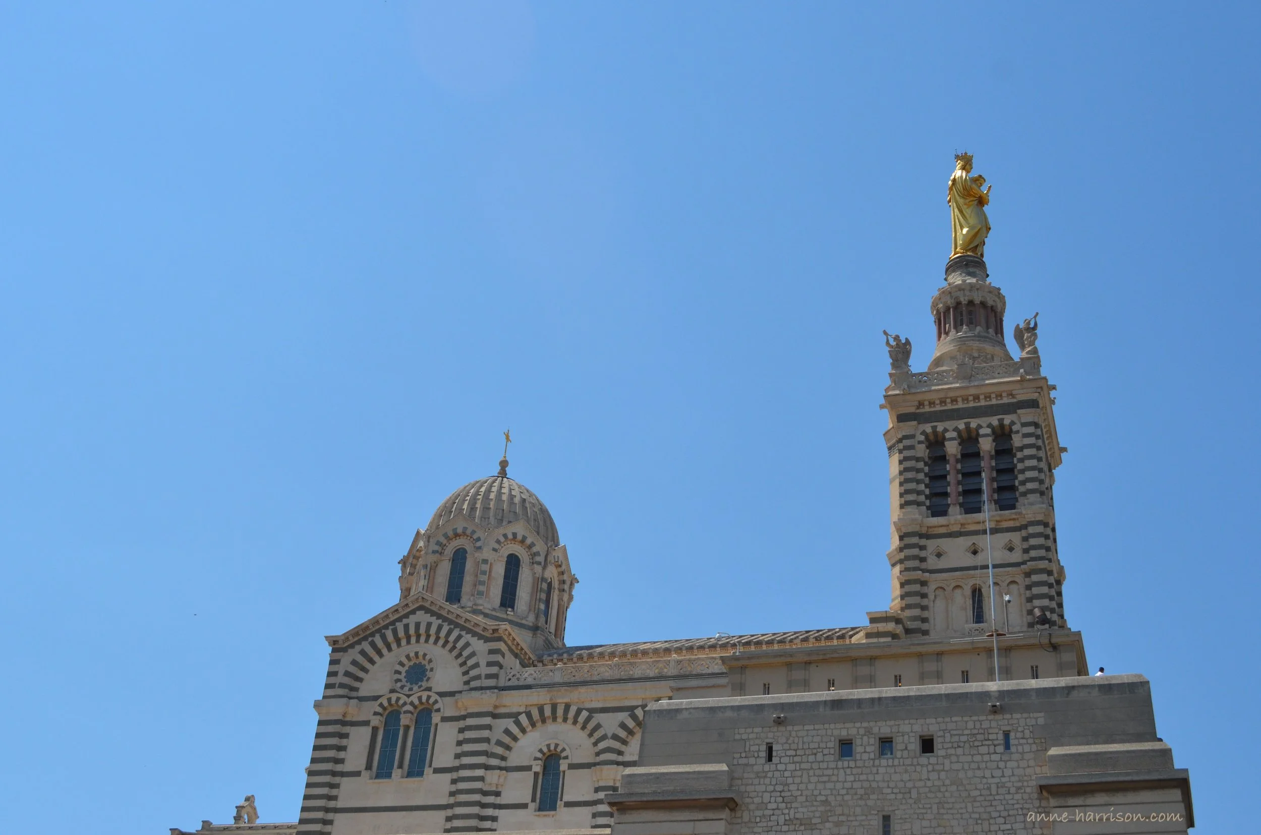 The exterior of the he Basilica de la Notre Dame de la Garde, showing the striped facade. The bell tower is topped with a golden madonna.