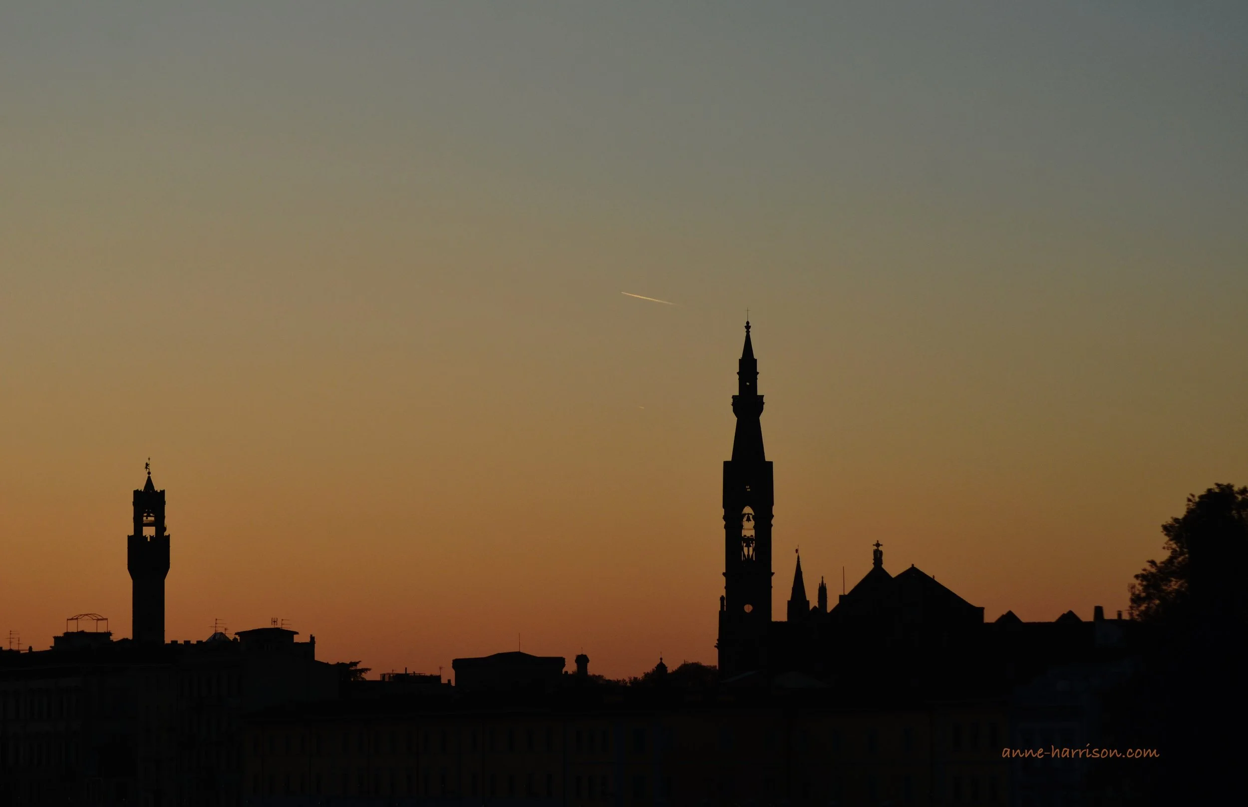 Towers in Florence are seen rising above the city at sunset. They are silhouetted against the sky.
