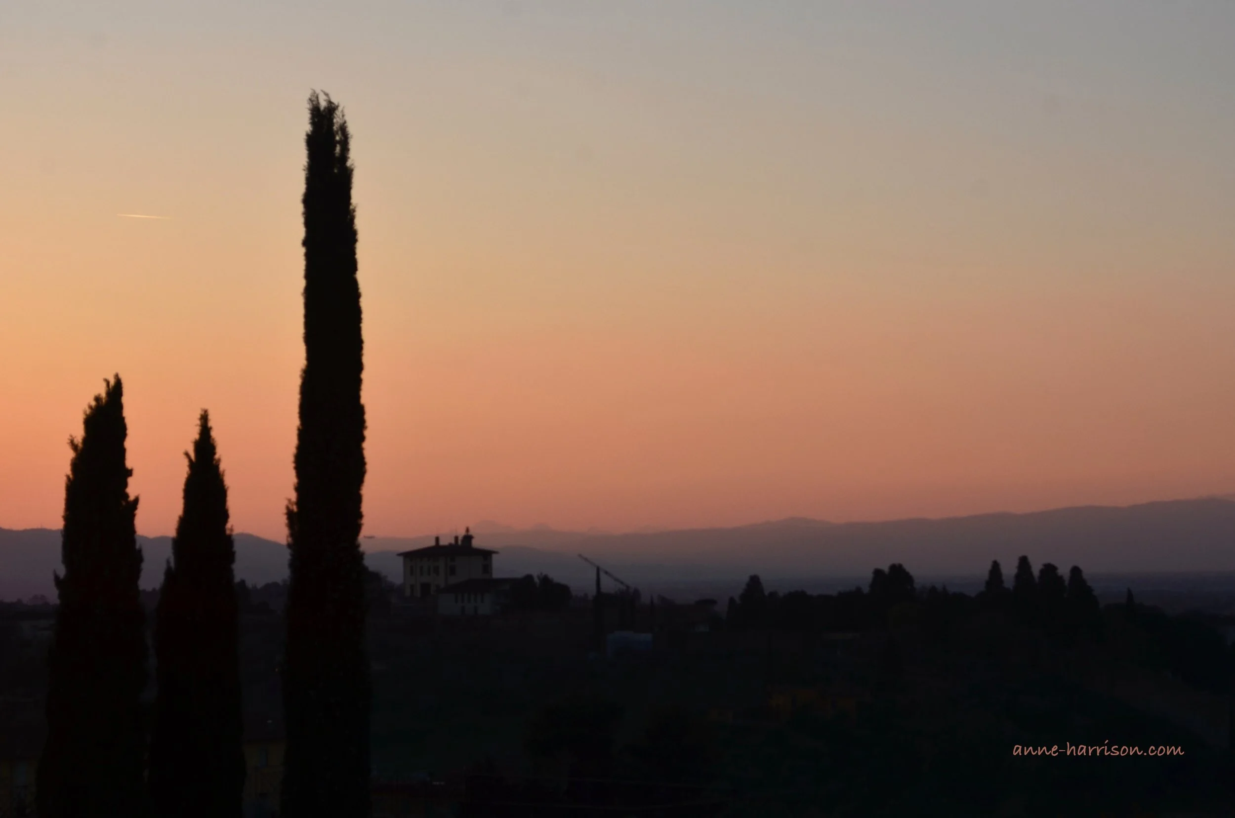 Cypress tower over a distant farmhouse in the Tuscan hills. The sky is the pinks and oranges of sunset, with the land almost purple. The trees are seen in silouhette against the sky.