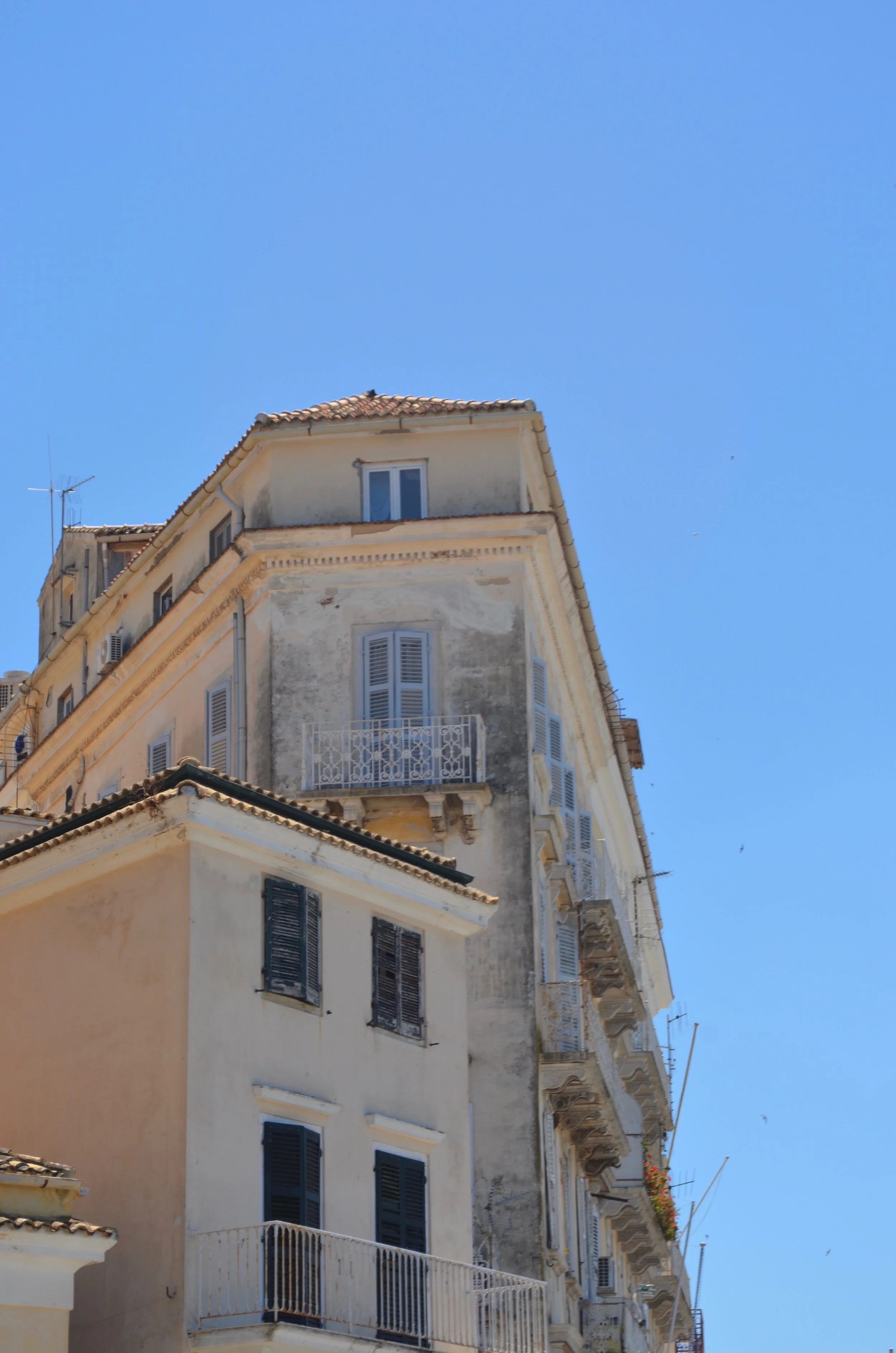  A weathered apartment block in Corfu. It is pale pink turning to grey. One window with blue shutters has a stone balcony with a white iron railing. 