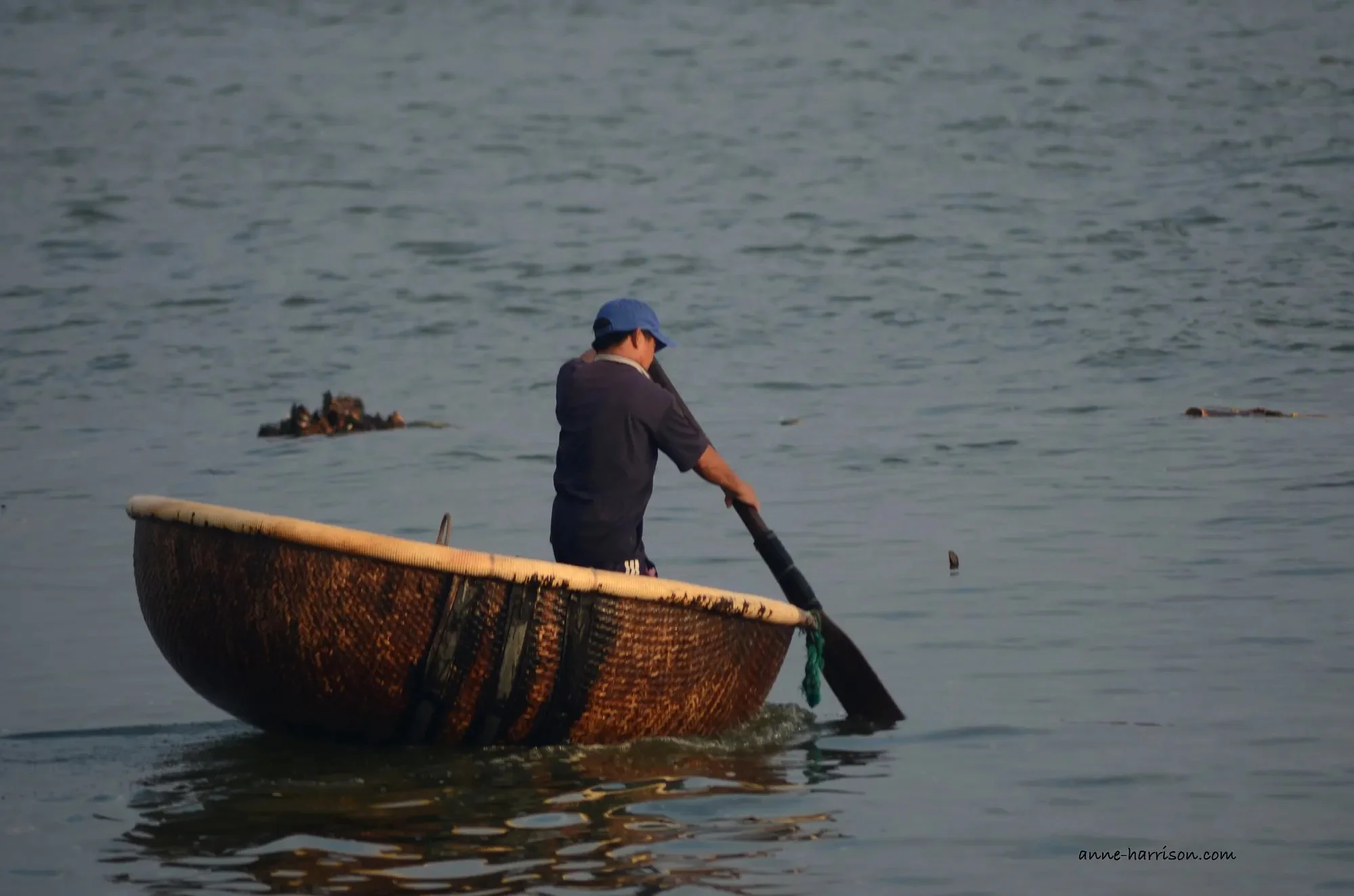 A man with a blue cap and dark blue shirt is steering a basket boat in still water.