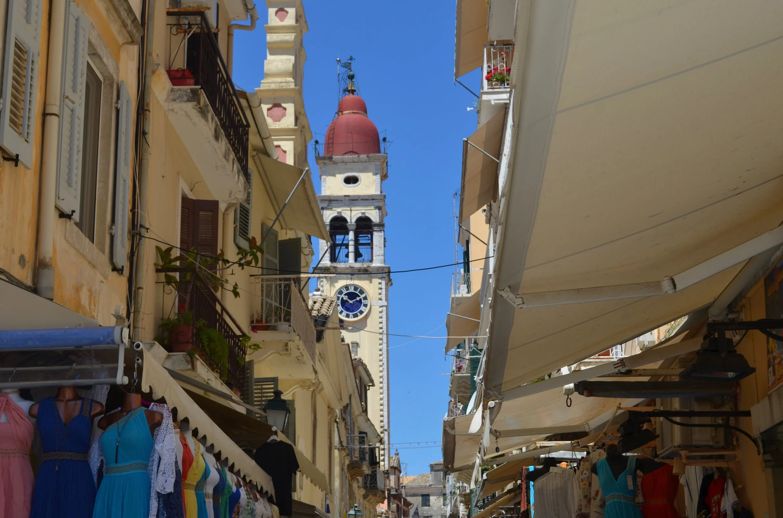 The bell tower of St Spyridon, Corfu, seen at the end of a street filled with shops. Clothes for sale hang beneath awnings outside the shops.