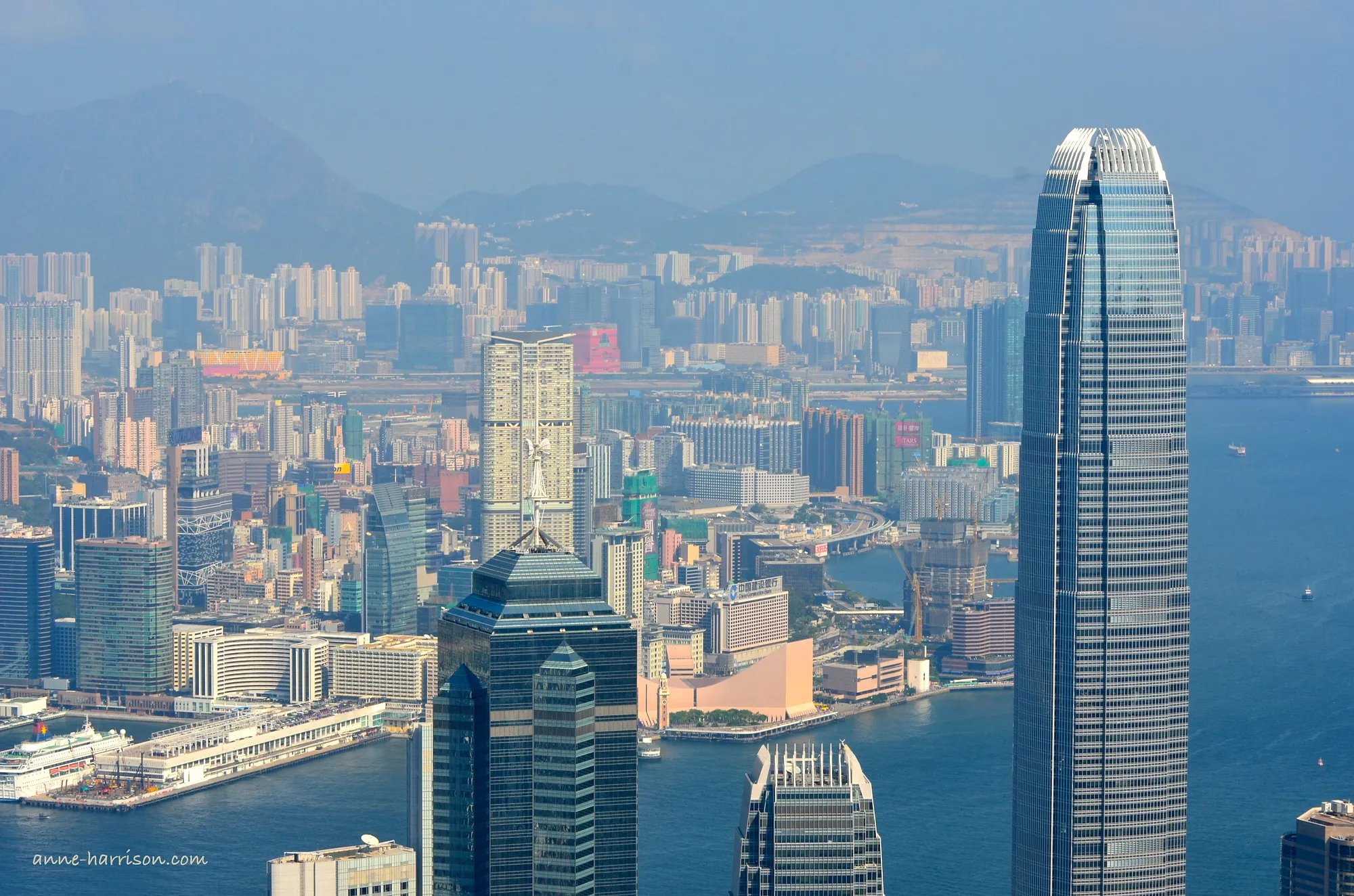 A view from The Peak down onto Hong Kong harbour and across to the International Cruise terminal and Kowloon. Tall office buildings fil the view, and mountains are just visible in the background.