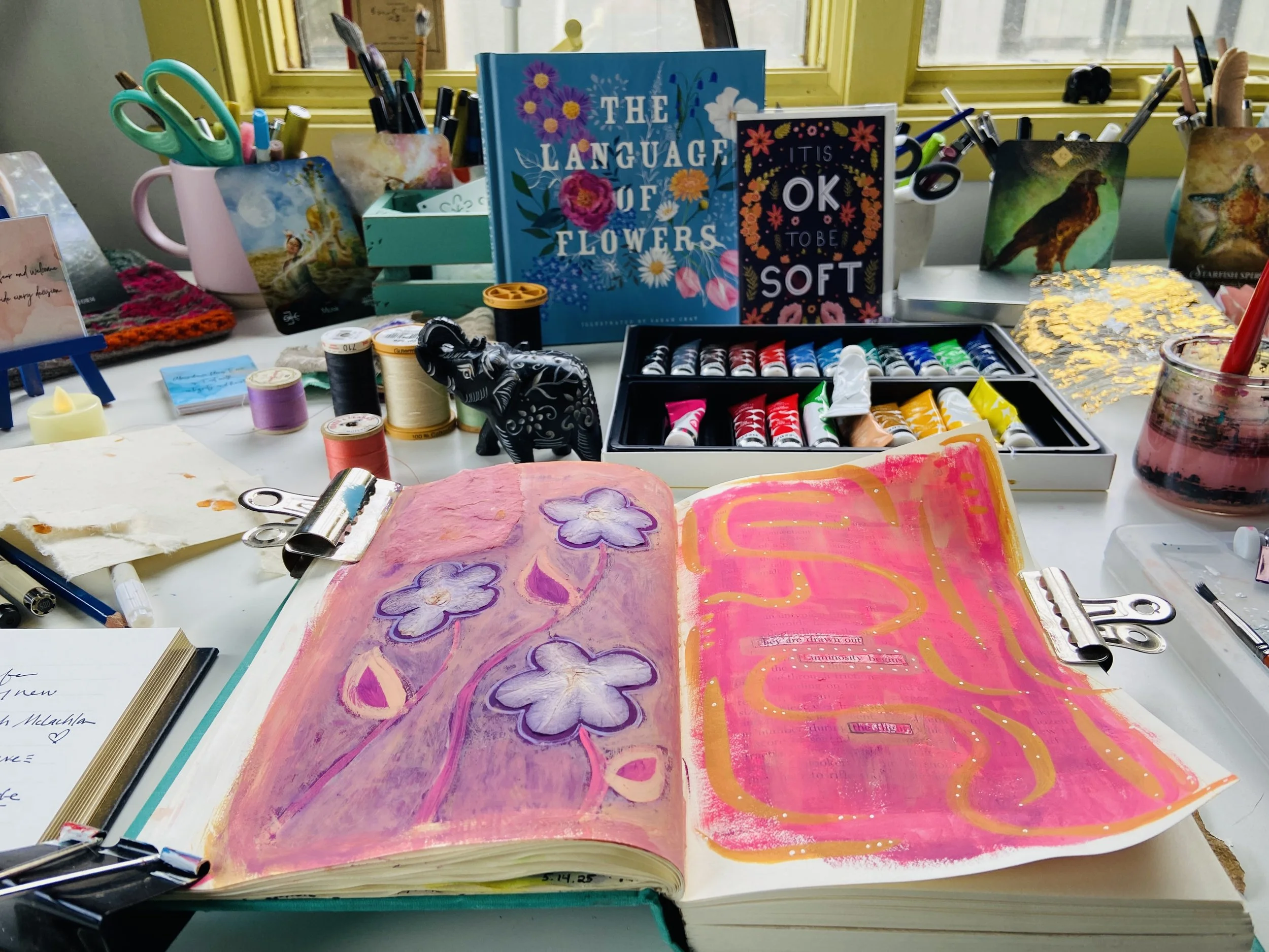 Emma Freeman's art studio table, art journal open with gathered purple flower petals & bright pink and purple gouache paint