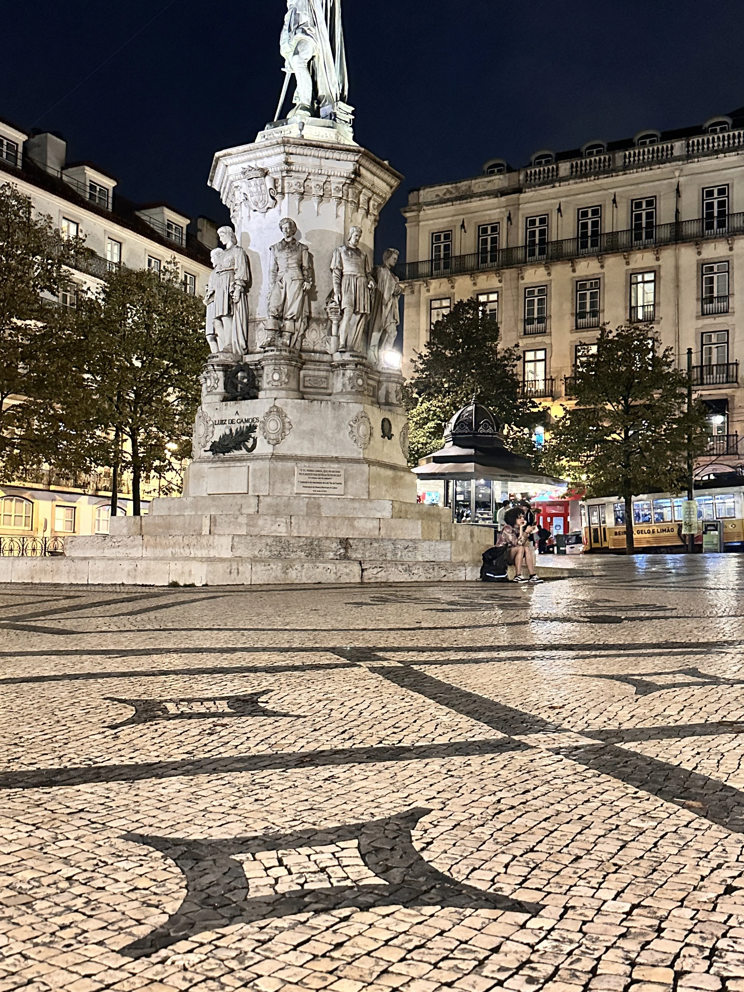 Lisbon Night view of a monument with statues on a tall pedestal, surrounded by trees and buildings, with a person sitting on the steps and a tram visible in the background.