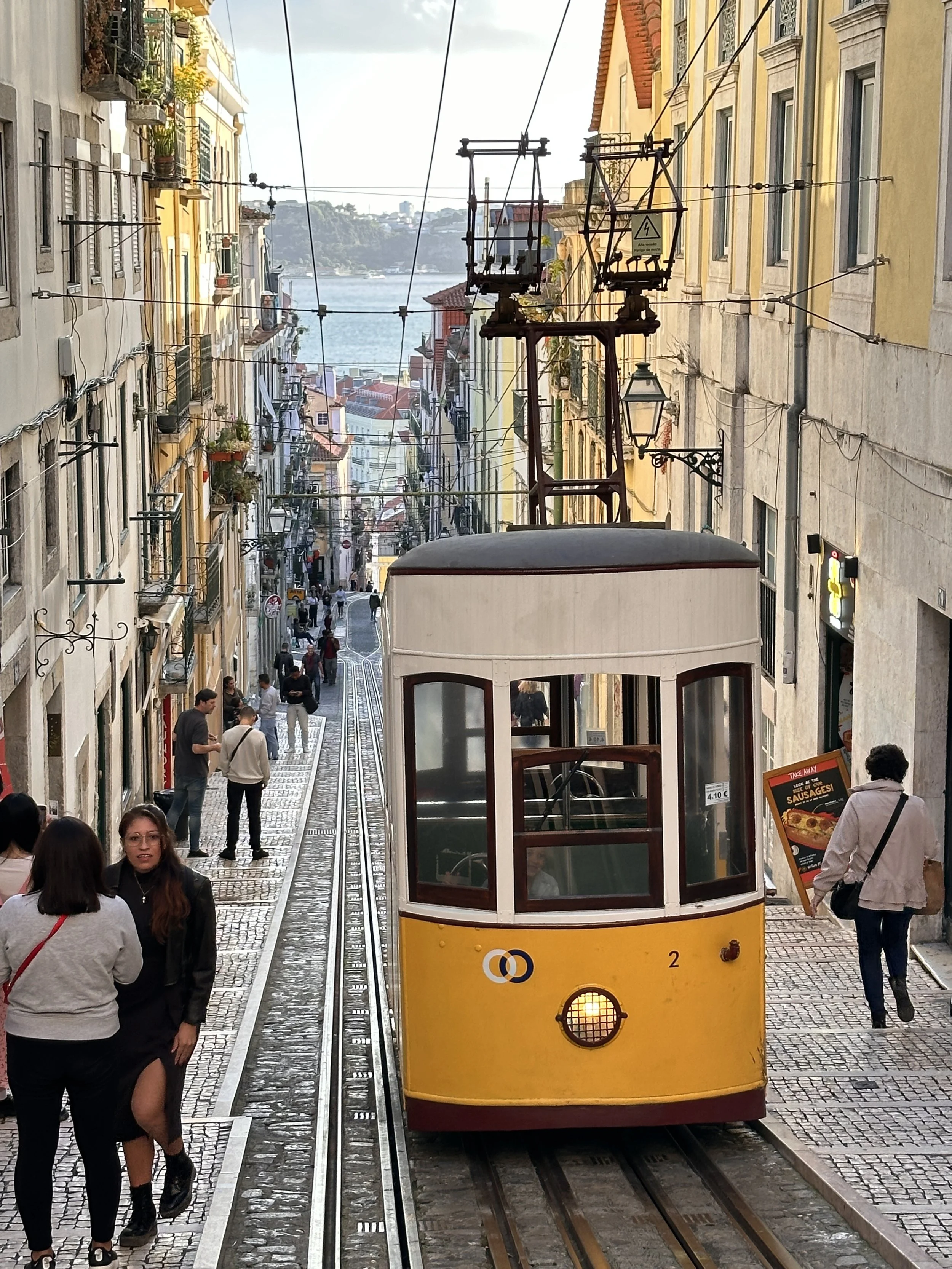 Don't Miss Lisbon Yellow and white tram moving through a cobblestone street lined with multi-story buildings, utility wires overhead, with pedestrians walking along the sidewalk.