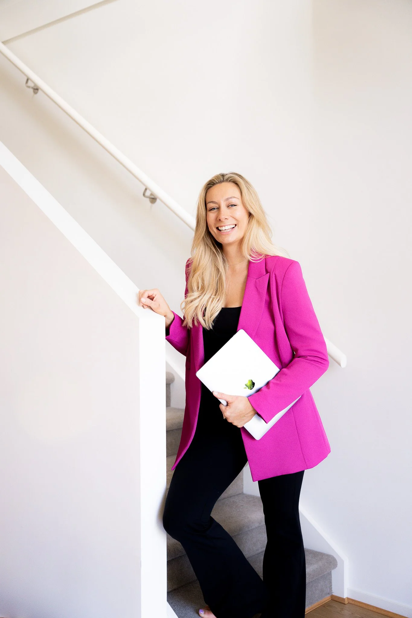 A woman with long blonde hair in a pink blazer and black pants holding a silver MacBook, smiling on a staircase inside a house.