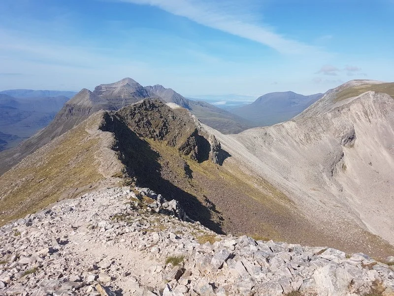 Mountain ridge with rocky terrain and distant mountain peaks under a partly cloudy sky.