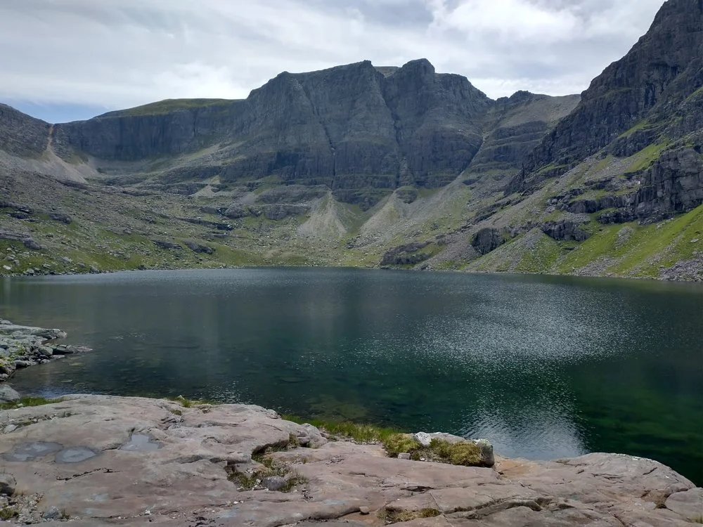 A serene mountain lake surrounded by rocky and grassy slopes with towering cliffs and cloudy sky above.