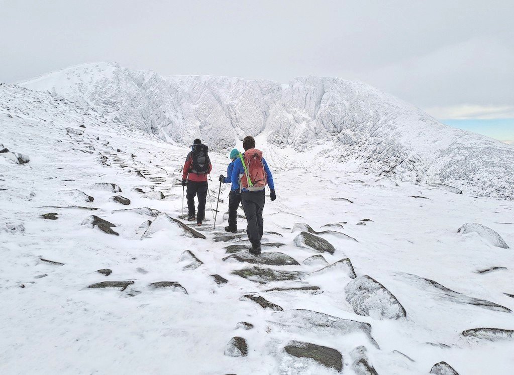 Three hikers in winter gear walking on a snow-covered mountain trail with rocks, approaching a snowy mountain ridge.
