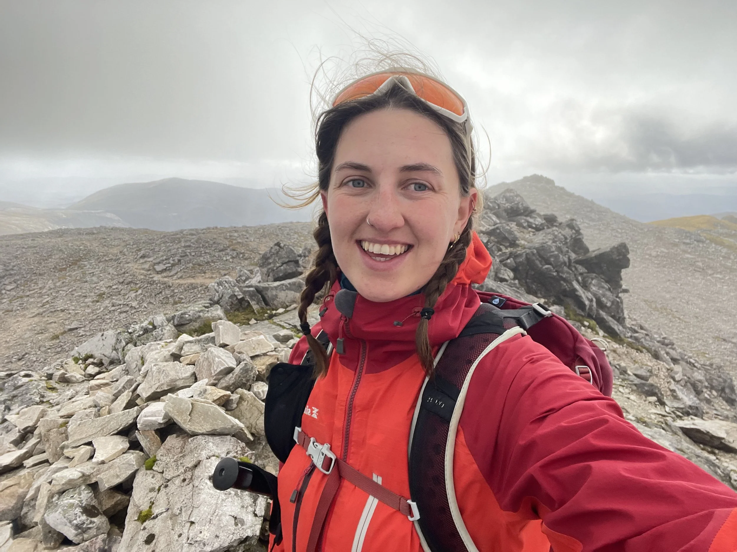 Woman in a red jacket with braids smiling on a rocky mountain trail, with cloudy sky and distant hills in the background.