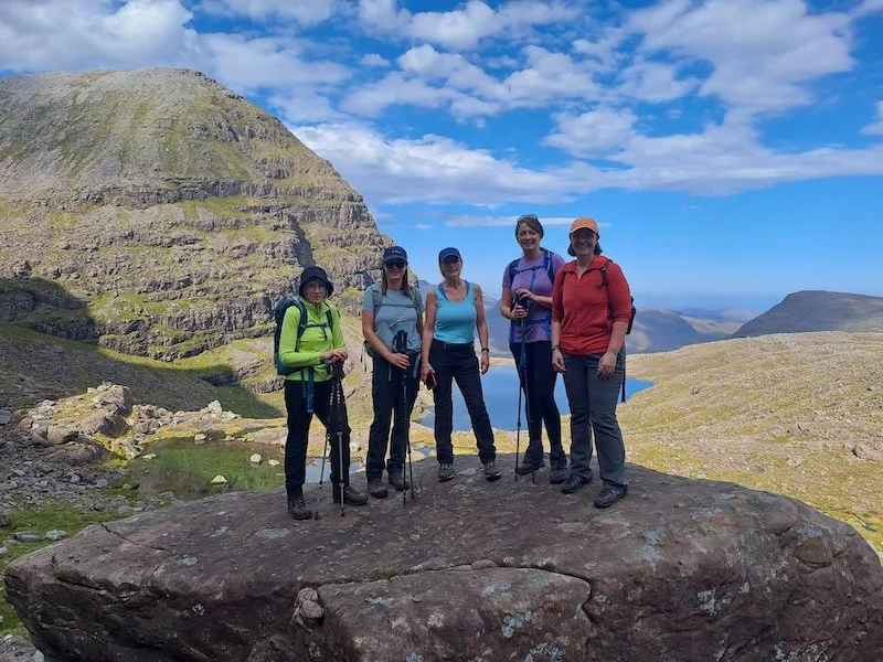 Five hikers standing on a large rock with a mountain and lake in the background under a partly cloudy sky.