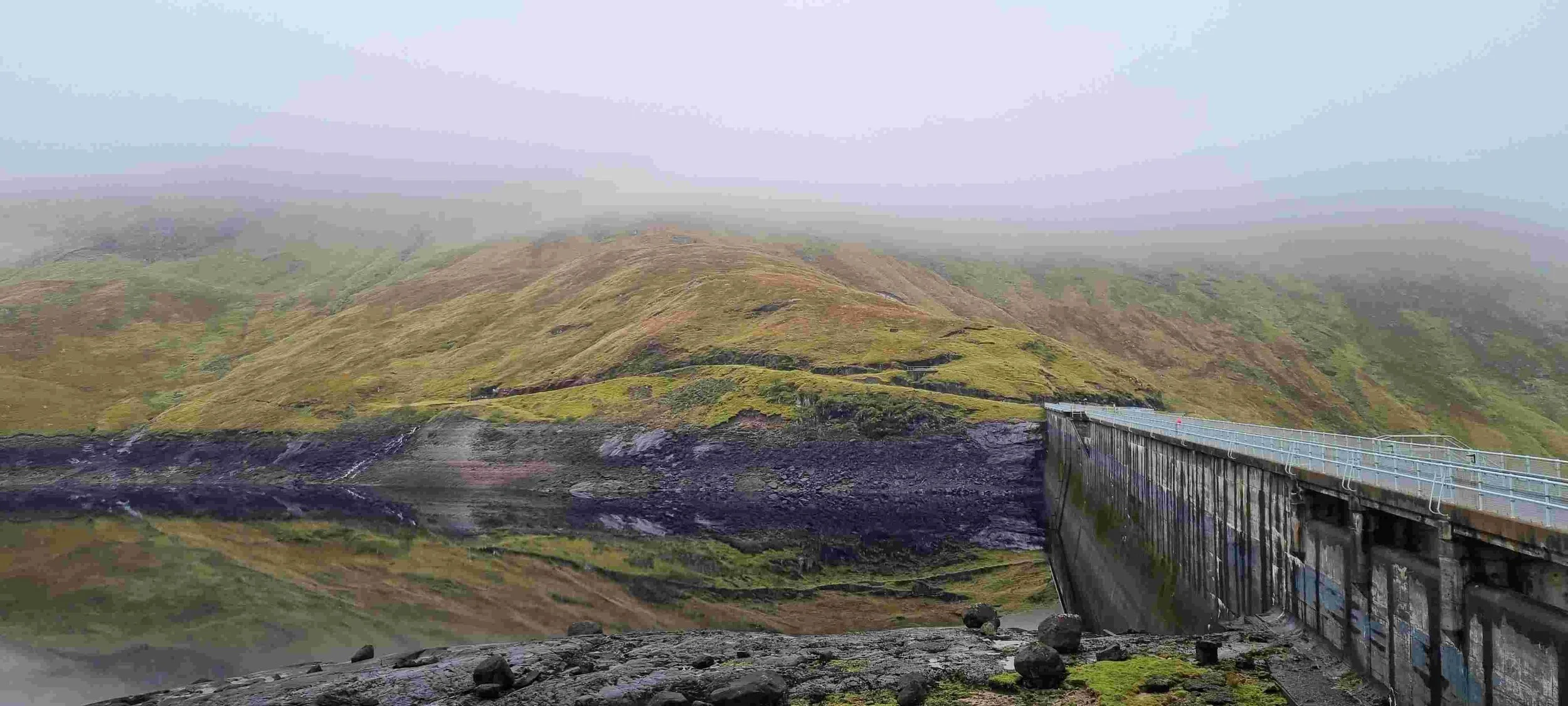 A bridge over a dark volcanic river in a foggy, mountainous landscape with green and brown hills.
