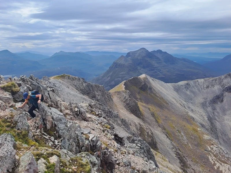 Hiker climbing rocky mountain terrain with mountains and cloudy sky in the background.