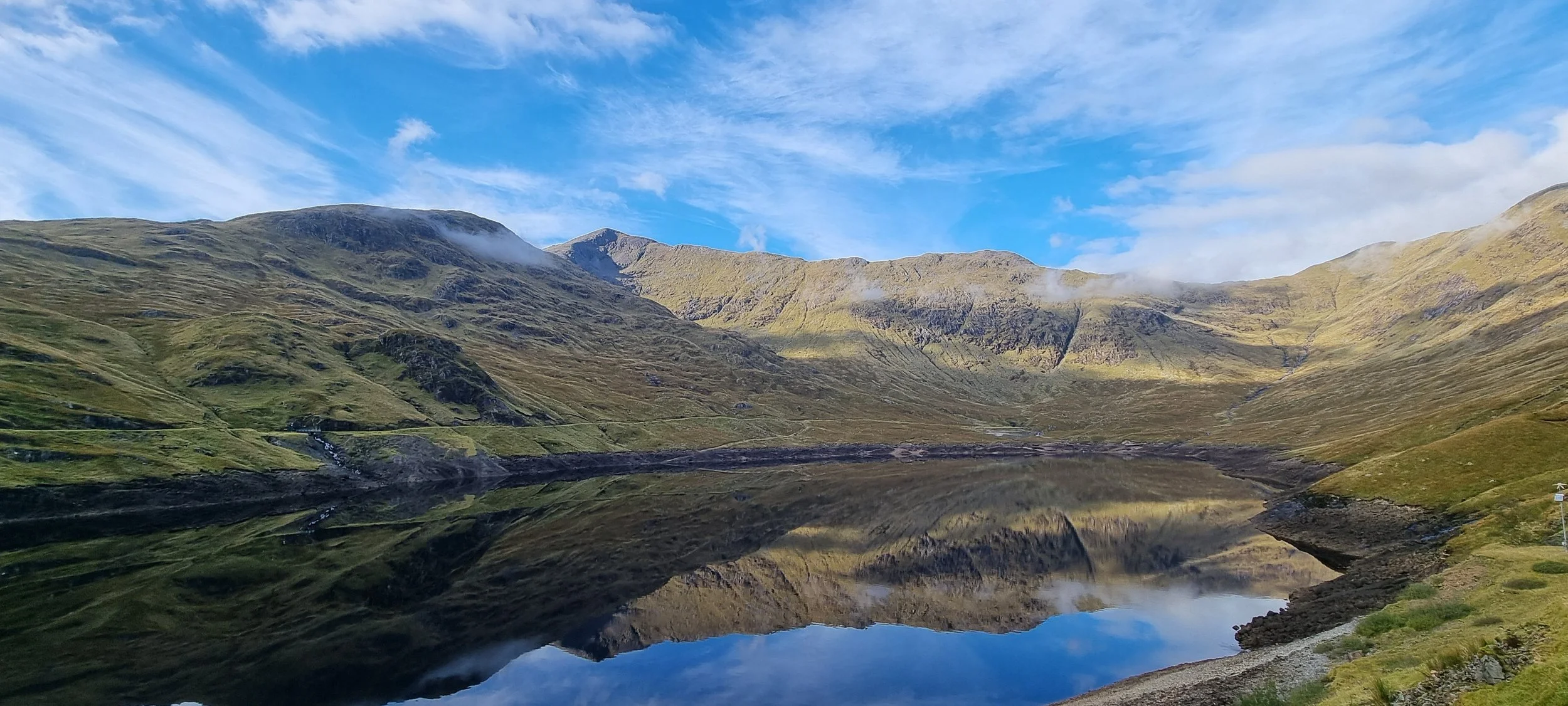 Scenic view of green mountains with a reflective lake in the foreground and a bright blue sky with scattered clouds overhead.