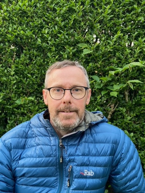 A middle-aged man with glasses and a beard wearing a blue Rab jacket standing outdoors in front of green leafy bushes.