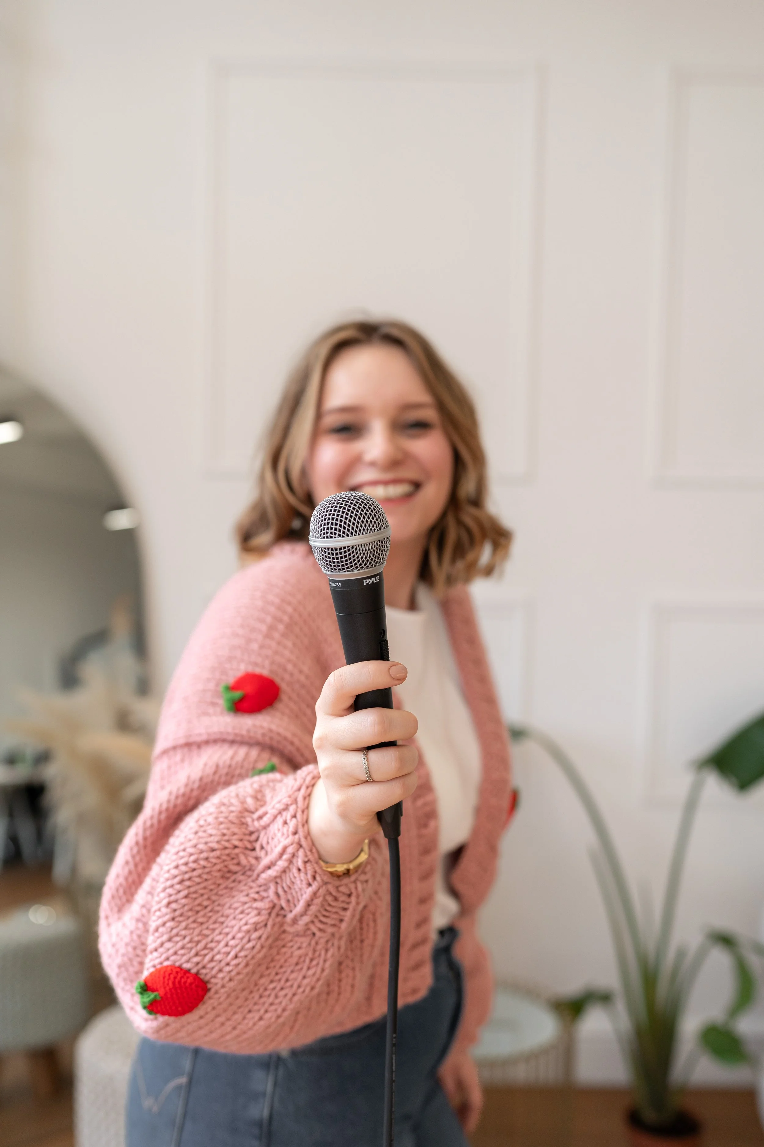 Smiling woman holding a microphone towards the camera, wearing a pink sweater with strawberry patches, in a cozy indoor setting with plants and furniture in the background.