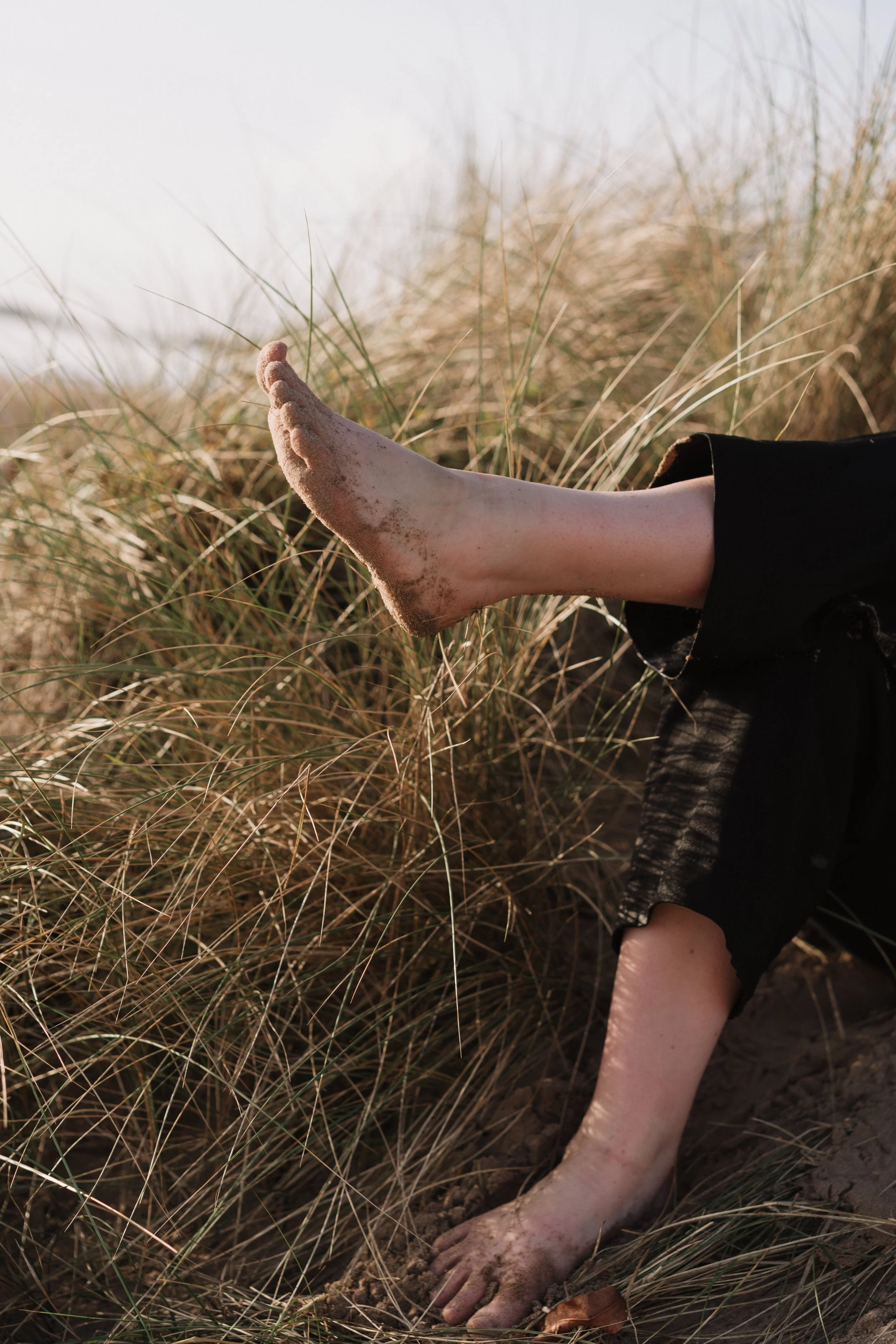 A person's legs and feet covered in sand, with one foot raised and resting against dry grass, wearing black pants rolled up at the ankles, outdoors on a sandy, grassy area.