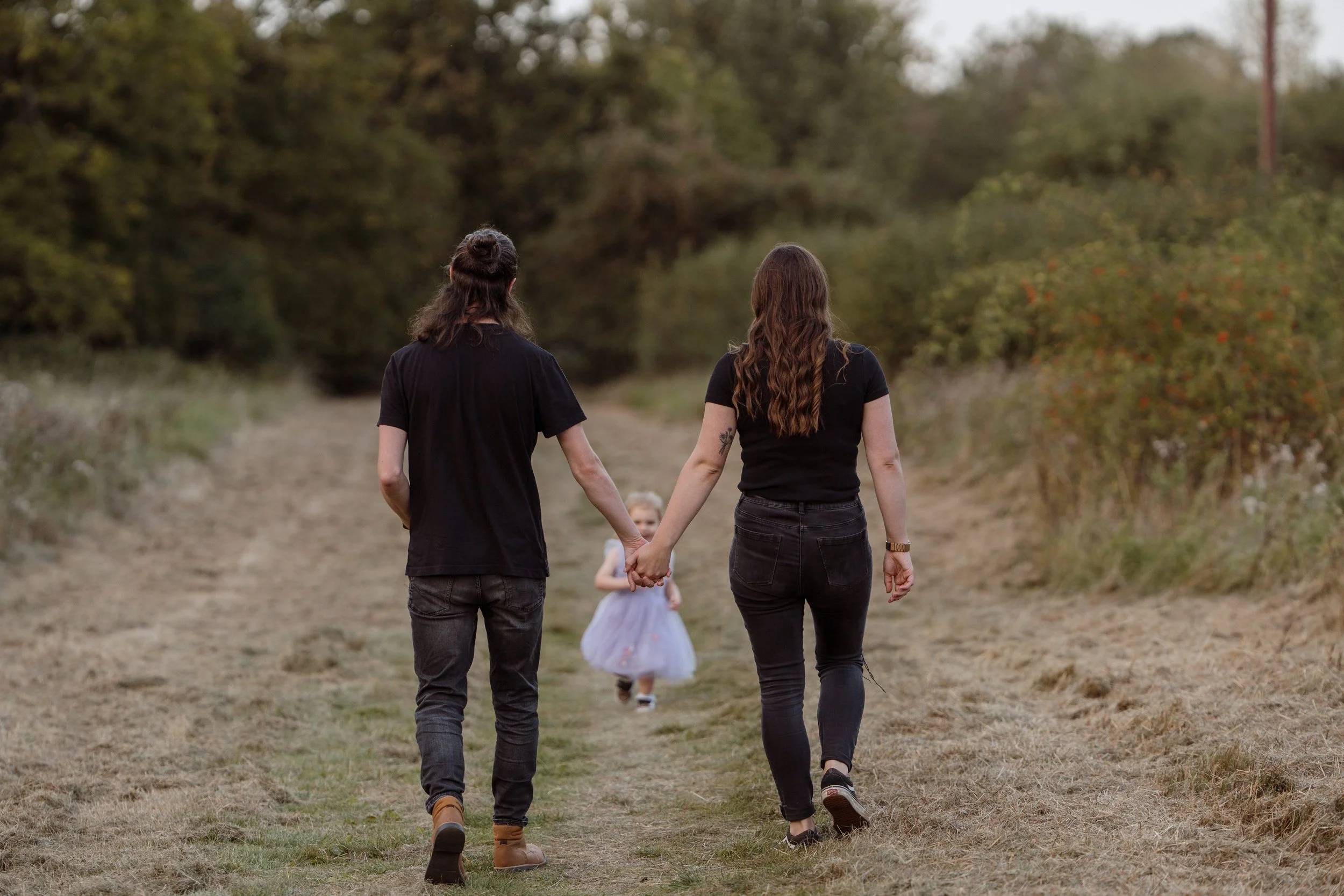 A couple walking hand in hand on a dirt trail with a young girl in a dress ahead of them in a natural outdoor setting.