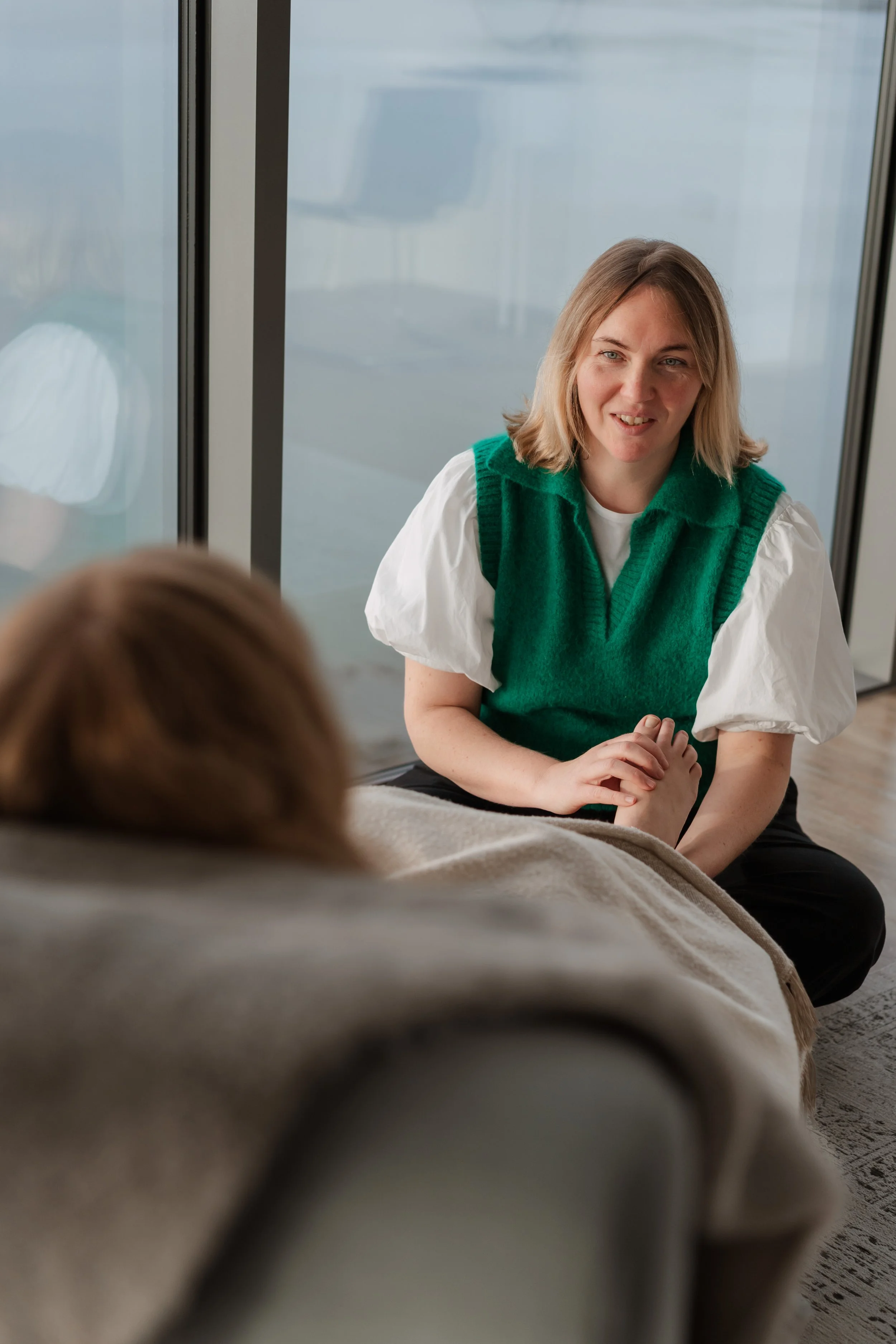 A woman with blonde hair wearing a green vest and a white puff-sleeve shirt sitting on the floor, holding hands with a person lying on a couch, near a large window.