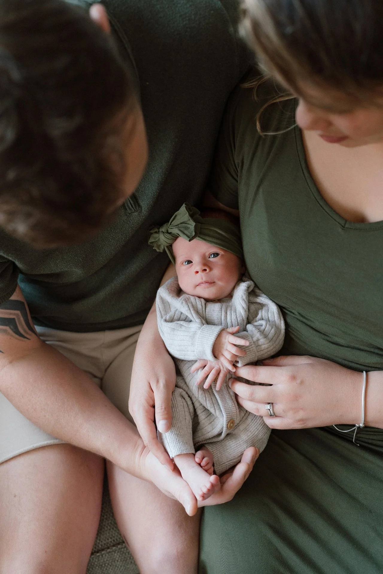 A newborn baby lying on a woman's chest, surrounded by two adults, one wearing an olive green top and the other a dark green top. The baby is wearing a beige sweater and matching pants, with a green headband and is looking directly at the camera.