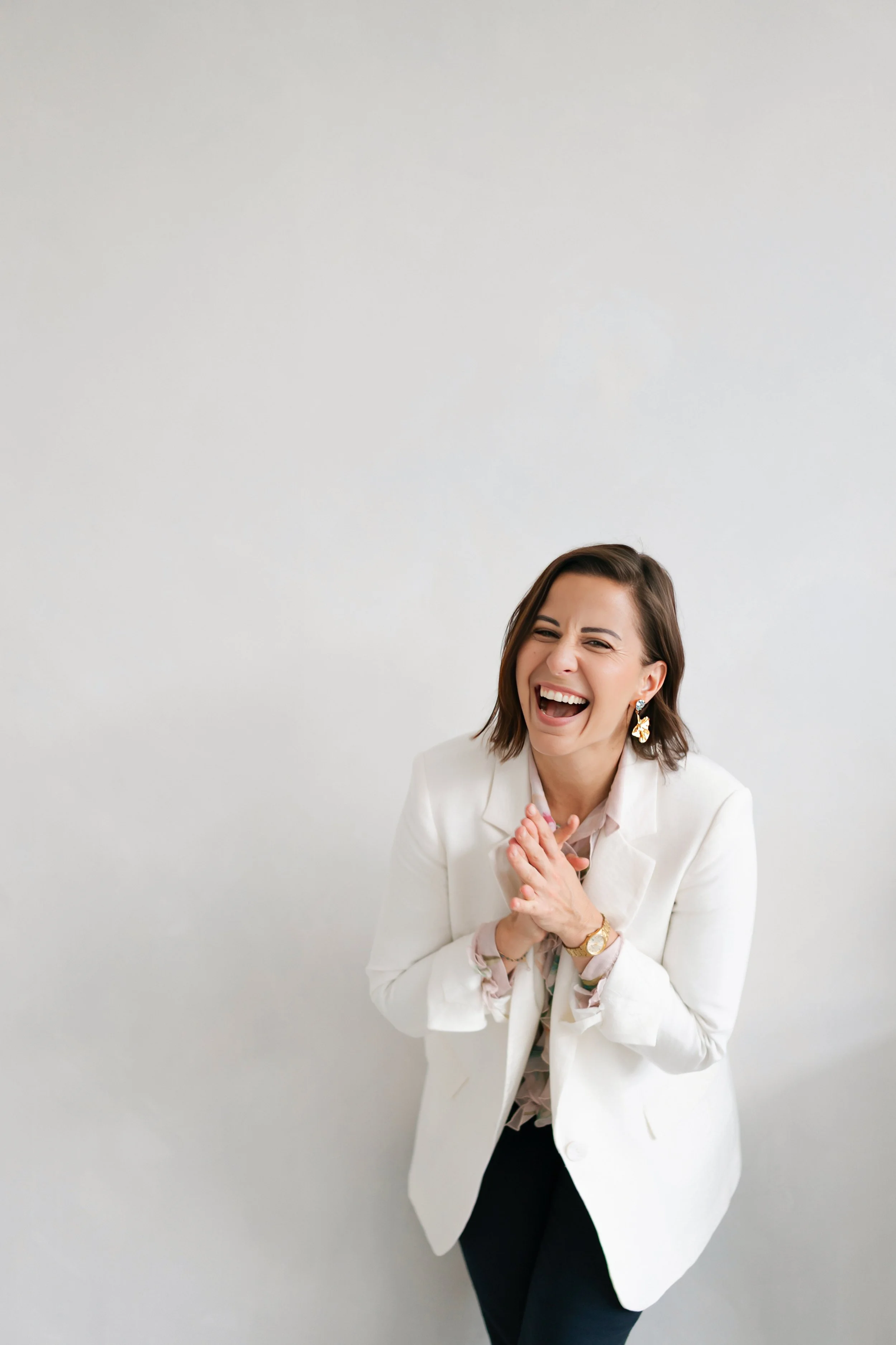 A woman with blonde hair smiling while leaning against a white wall, holding a camera with a large lens in a bright, modern room with a potted plant and a white chair in the background.