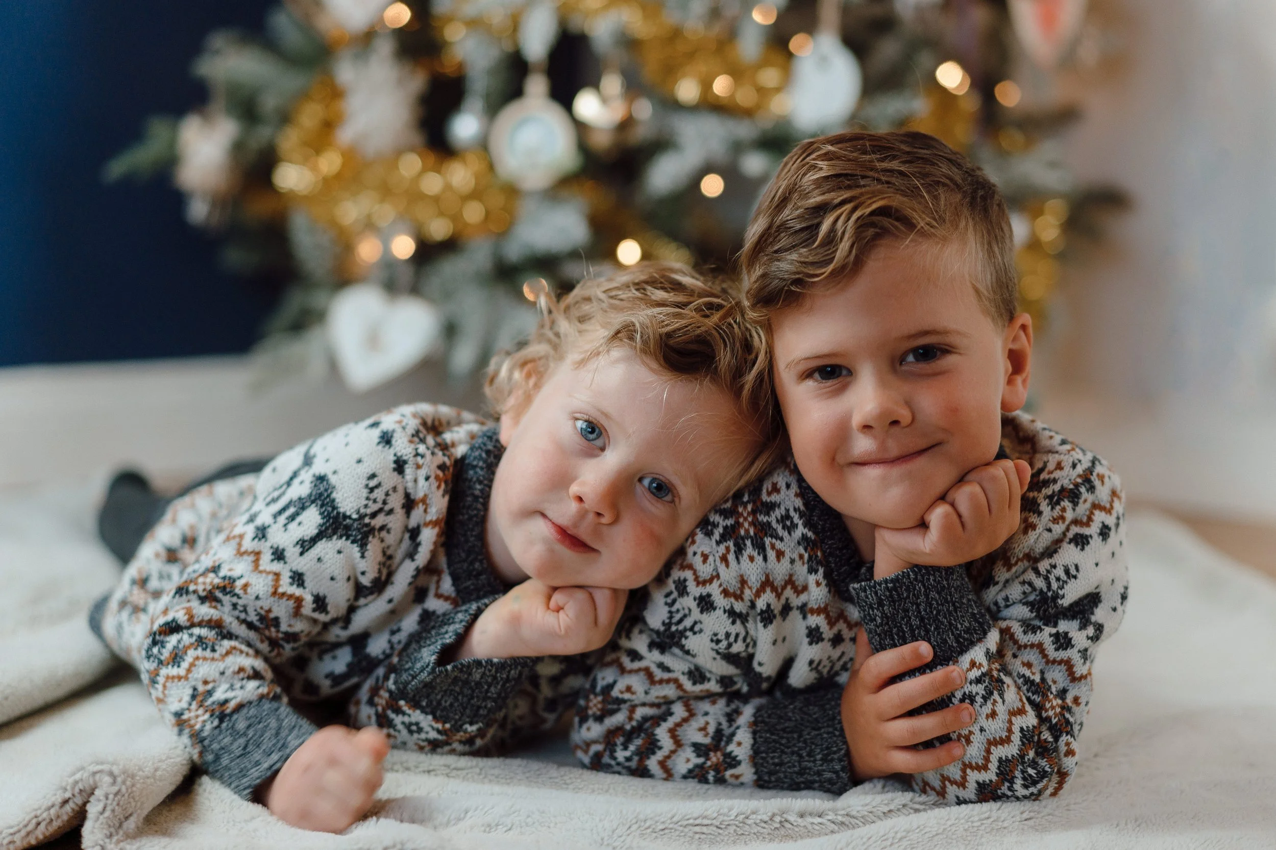 Two young children, a girl with blue eyes and wavy blonde hair and a boy with brown hair, are lying on a soft blanket in front of a decorated Christmas tree, wearing matching Christmas sweaters, with the girl resting her head on her hand and the boy 