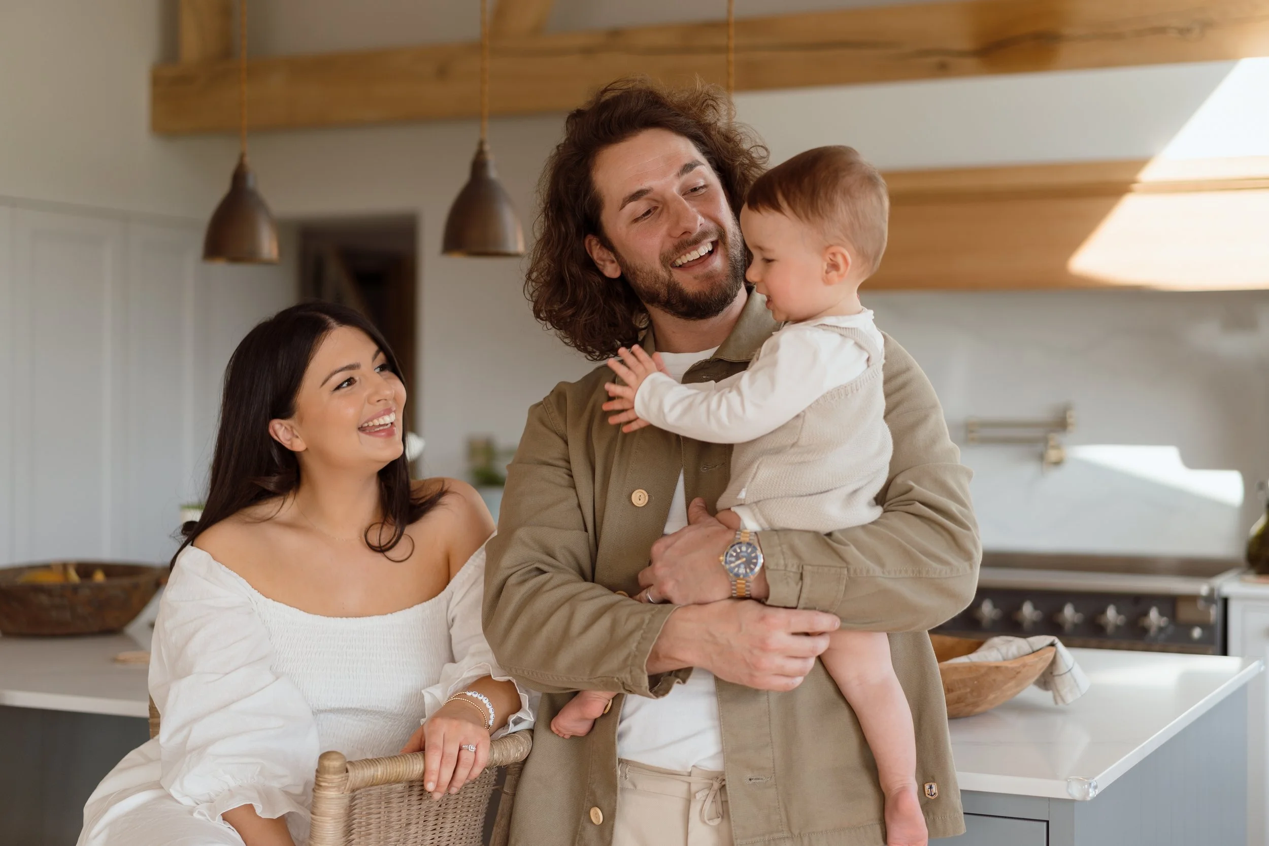 A happy family in a bright kitchen, with a man holding a young child, a woman smiling and sitting nearby, all sharing a joyful moment.