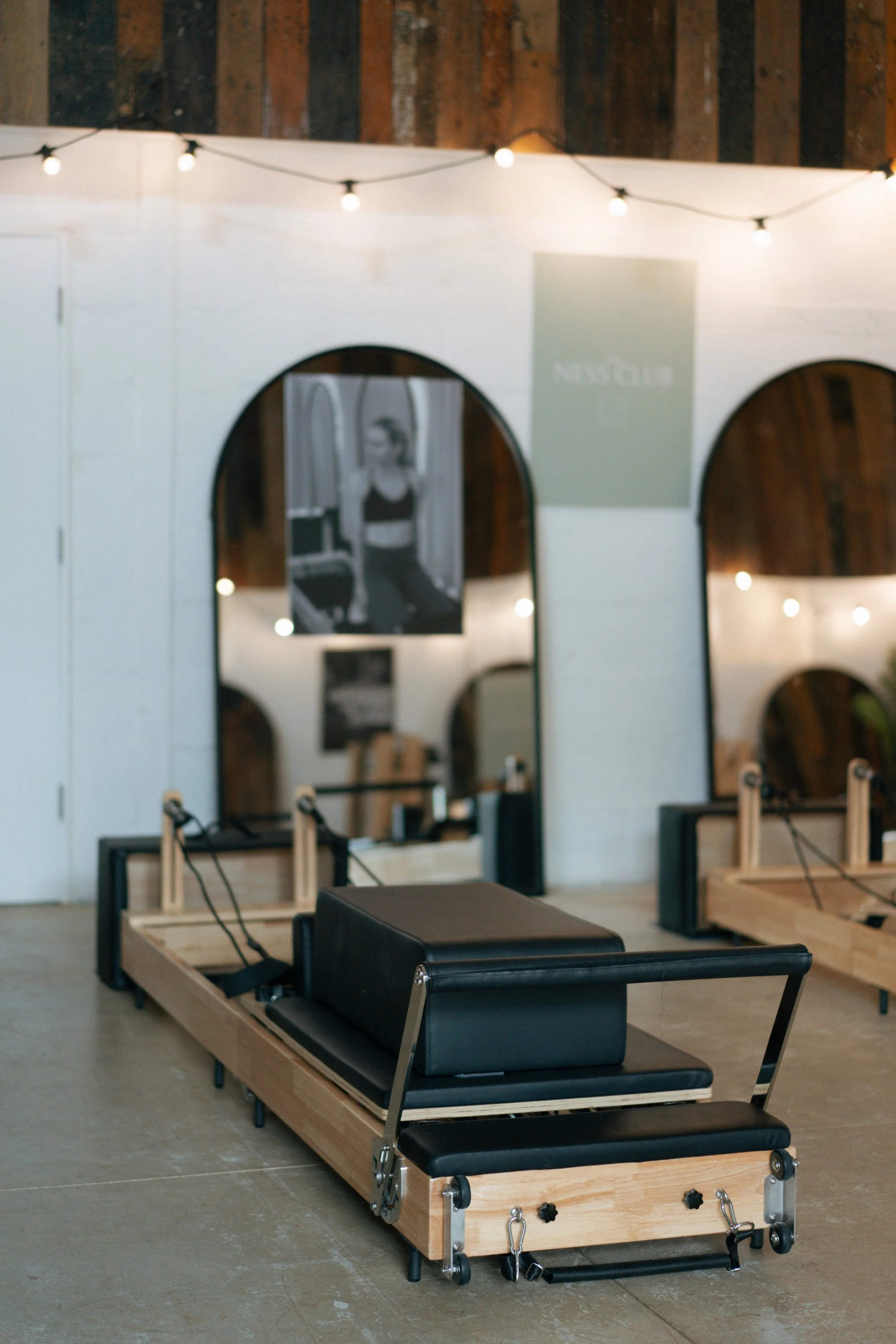 A Pilates reformer machine in a fitness studio with mirrors and string lights on the wall.