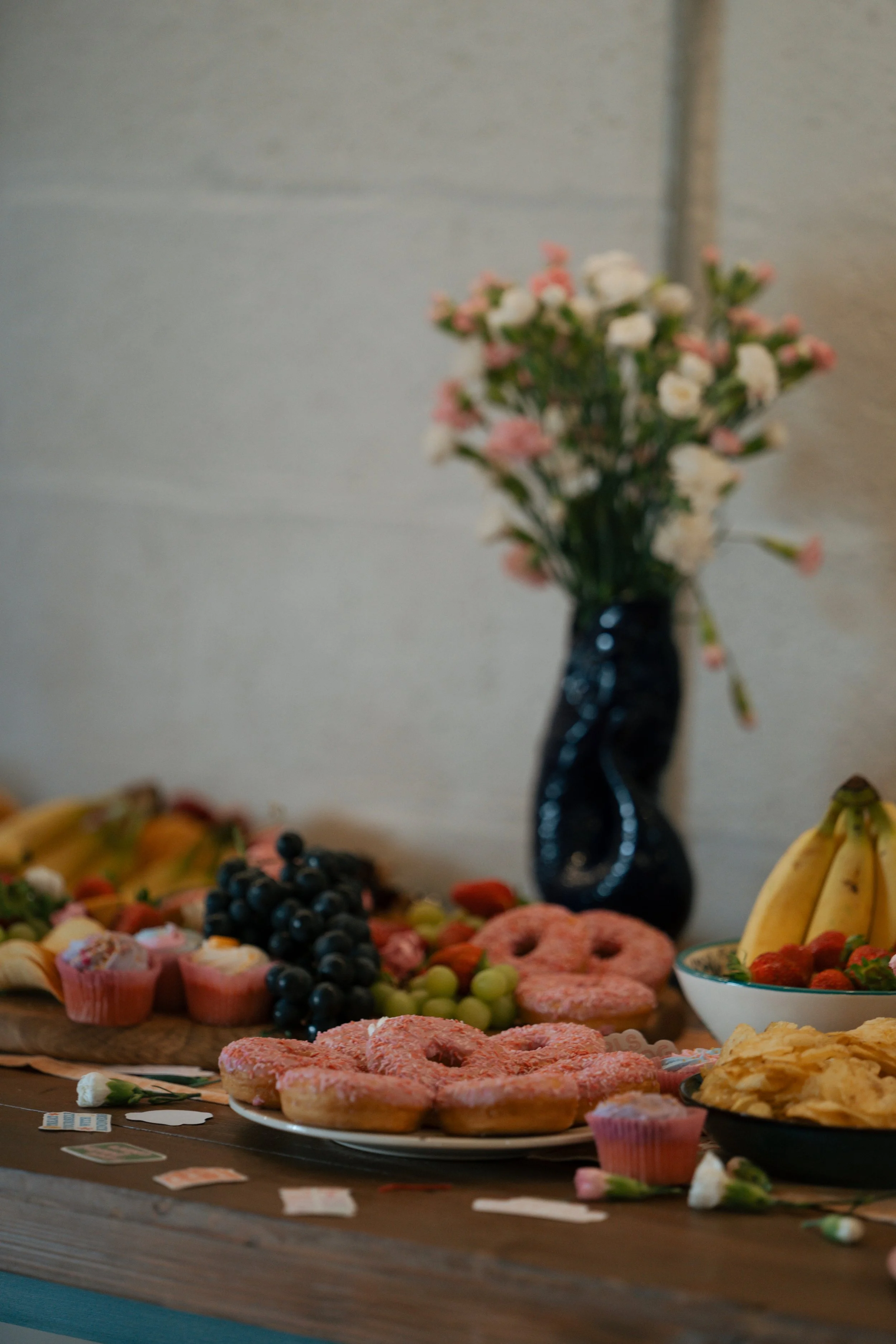 A table with pink frosted donuts, grapes, strawberries, bananas, cupcakes, chips, and a glass vase with pink and white flowers.