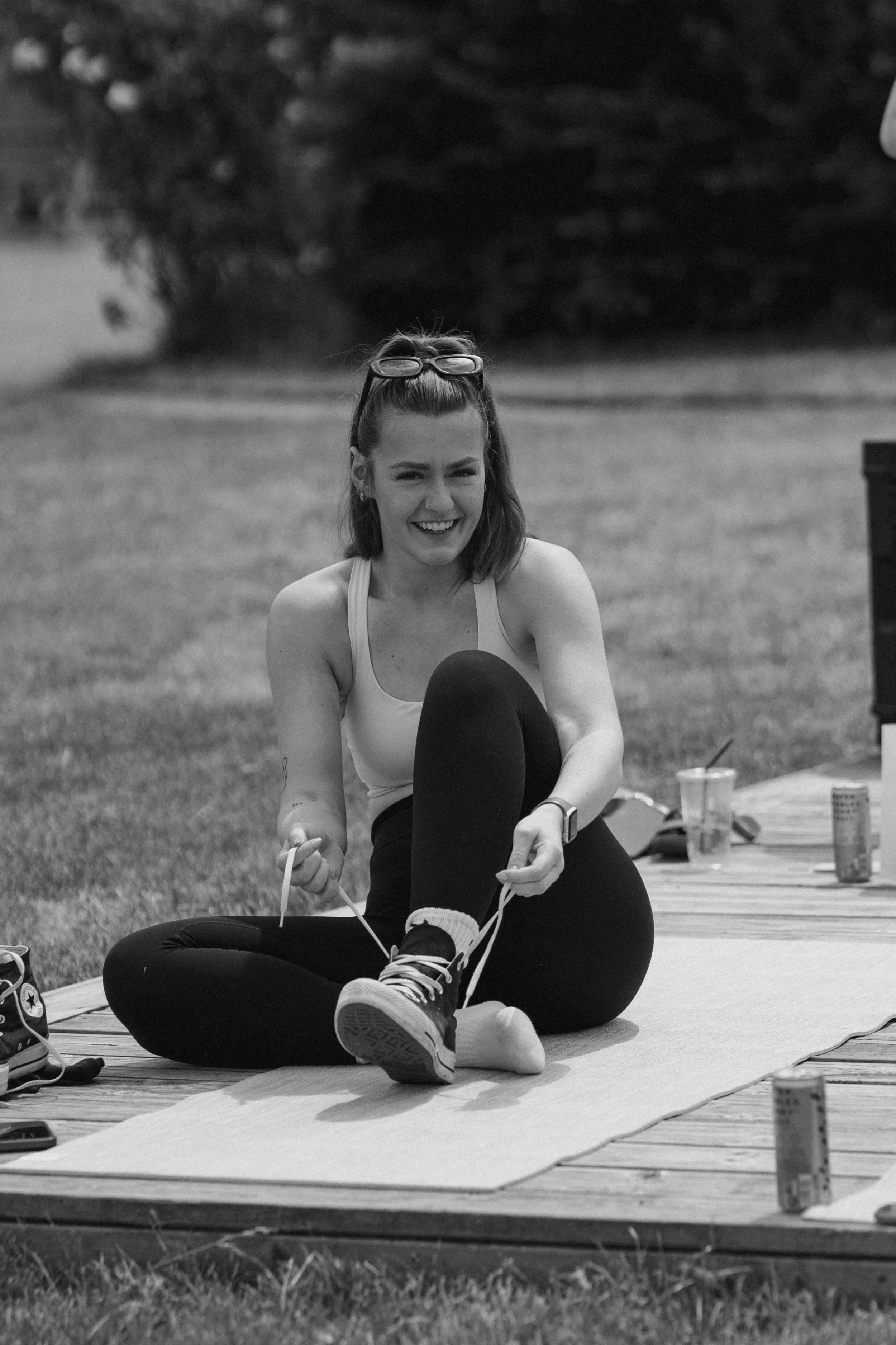 A woman sitting outdoors on a mat, tying her shoelaces, smiling, with sunglasses on her head, surrounded by drinks and snacks.