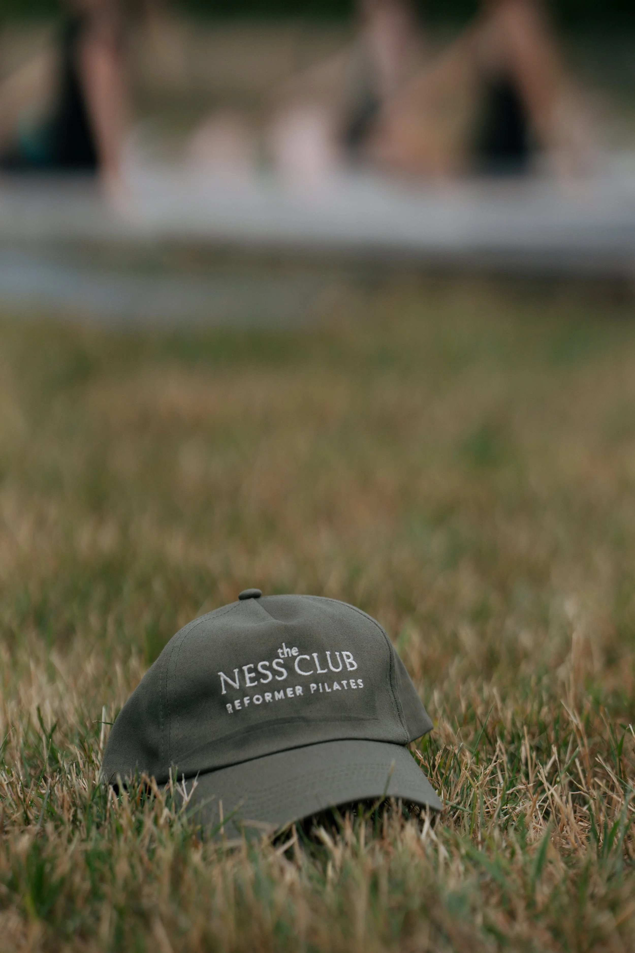 A gray baseball cap on grass with the text 'the NESS CLUB REFORMER PILATES' embroidered on the front.