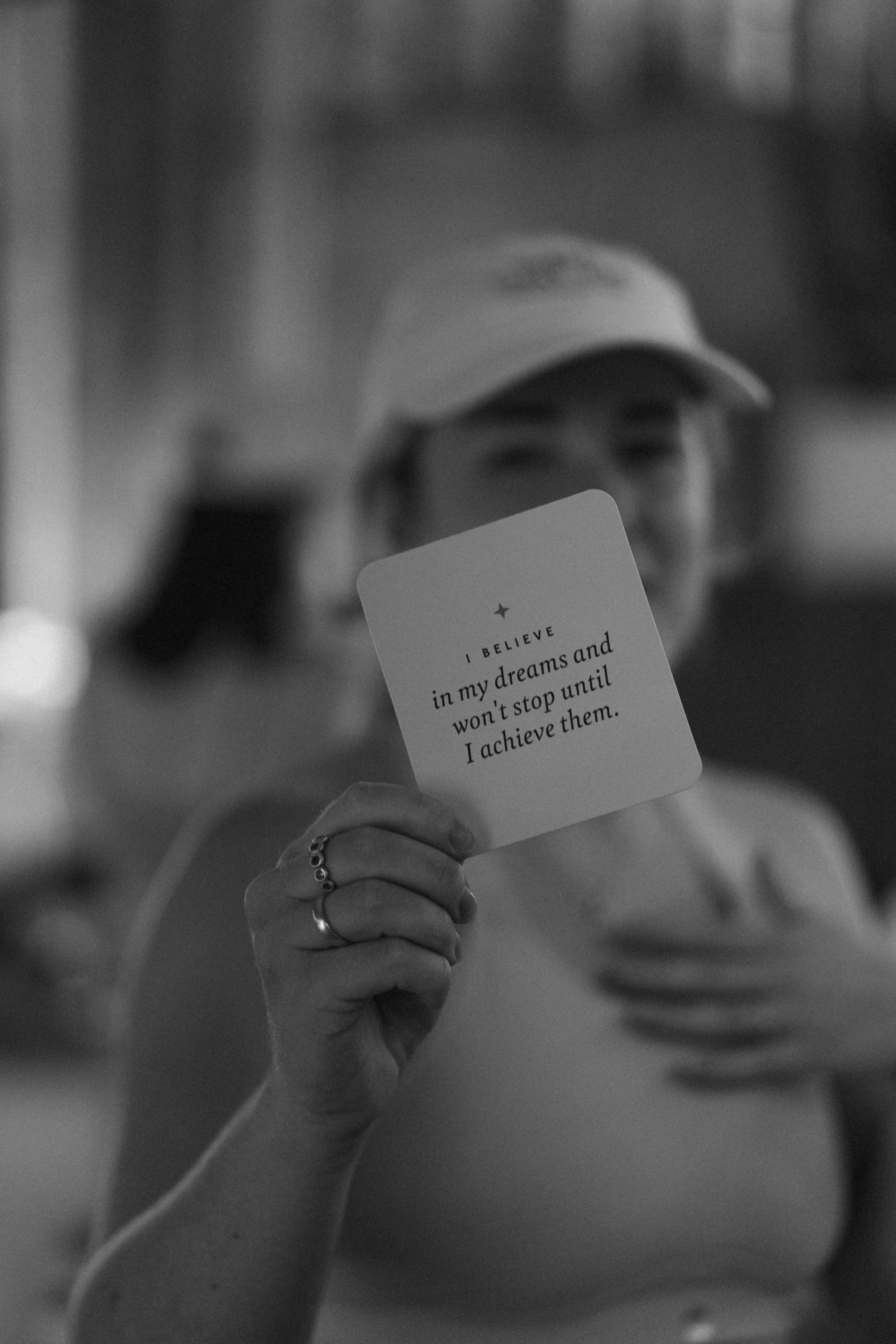 A woman wearing a hat holding a card with an inspiring quote, with her hand raised towards the camera. The quote reads, "I BELIEVE in my dreams and won't stop until I achieve them."