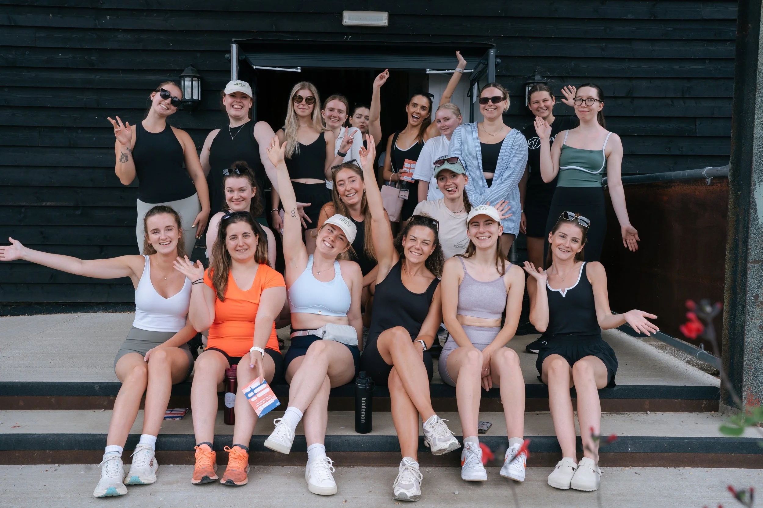 Group of nineteen young women smiling and posing in front of a black wooden wall, some sitting on stairs and others standing, during a daytime outdoor event.