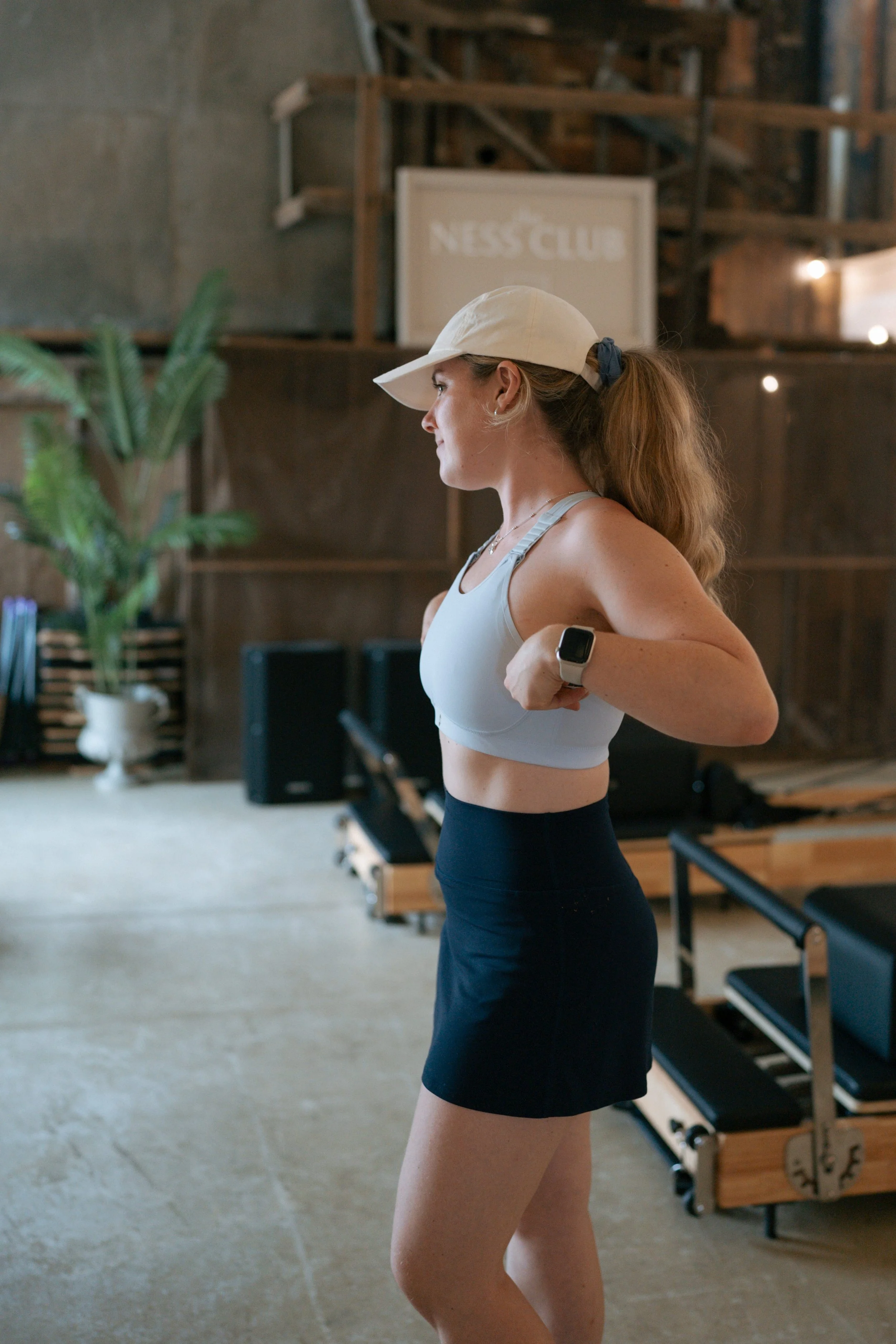 A woman in workout attire, including a light gray sports bra, black shorts, a beige baseball cap, and a smartwatch, is preparing for exercise in a gym with wooden and concrete decor, with a plant and exercise equipment in the background.