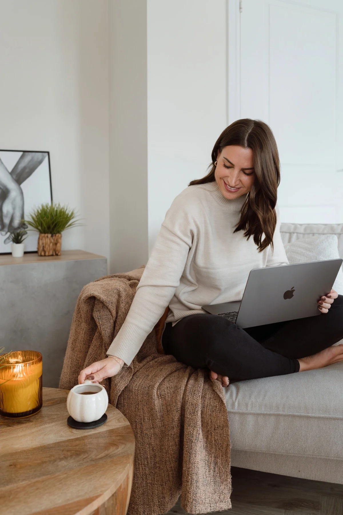 A woman sitting on a couch with a laptop, reaching for a coffee mug, smiling in a cozy living room.