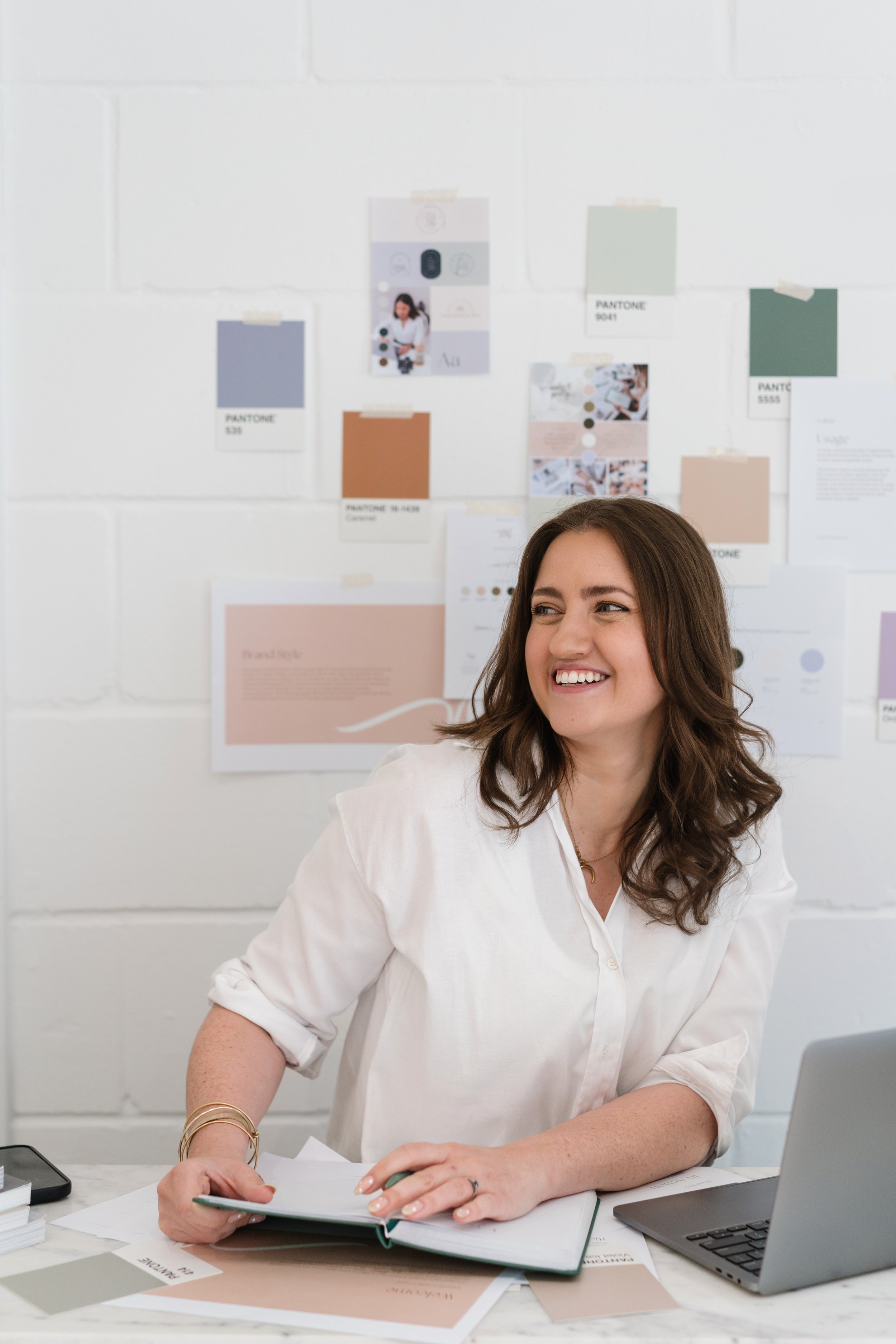 A brand photographer capturing a designer at work - smiling woman seated with her laptop surrounded by colour swatches and mood board inspiration, natural brand photography by Photography by Trish, Cambridge.
