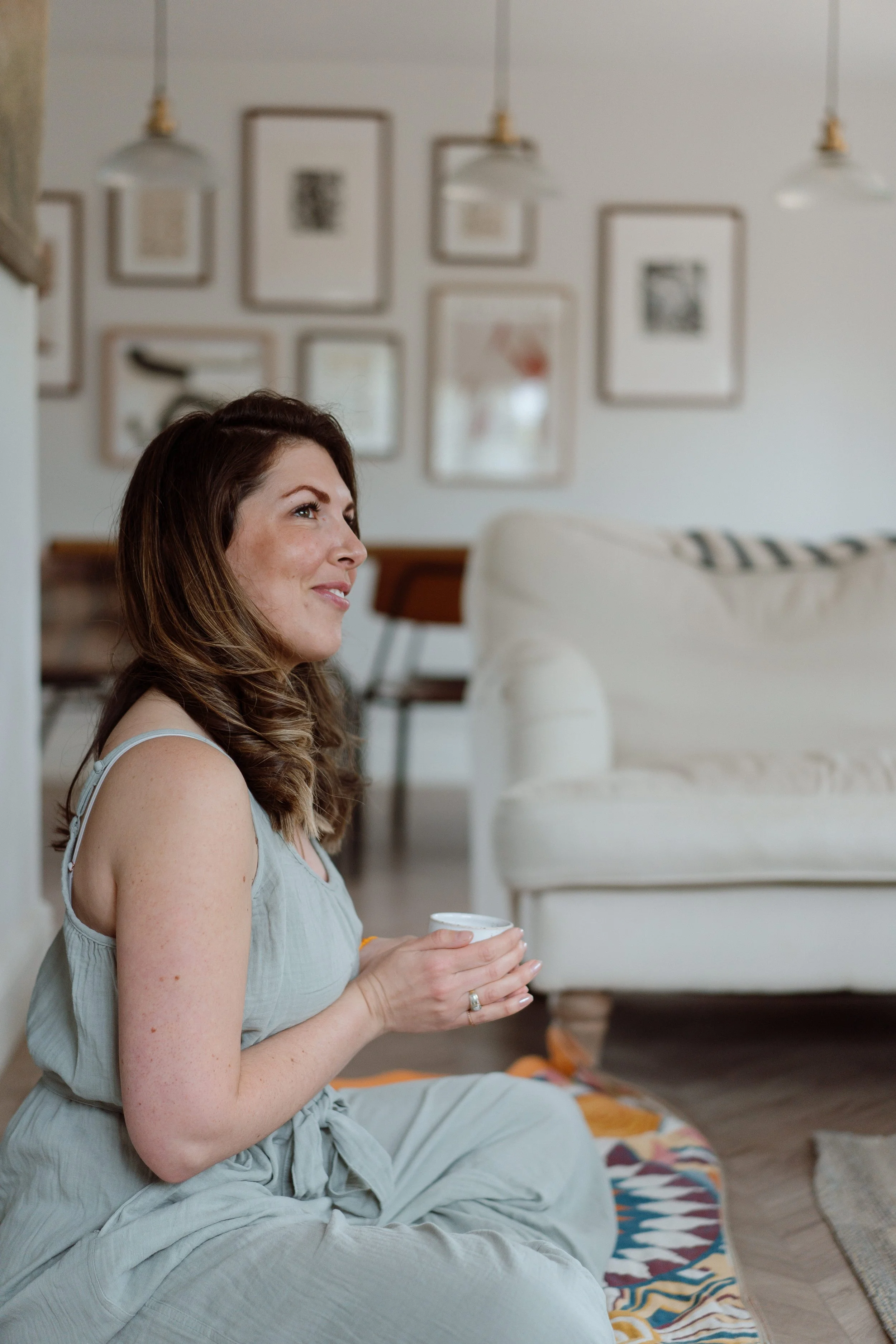 A woman with brown hair sitting cross-legged on a colorful rug, holding a white mug, smiling and looking to the side in a cozy living room.