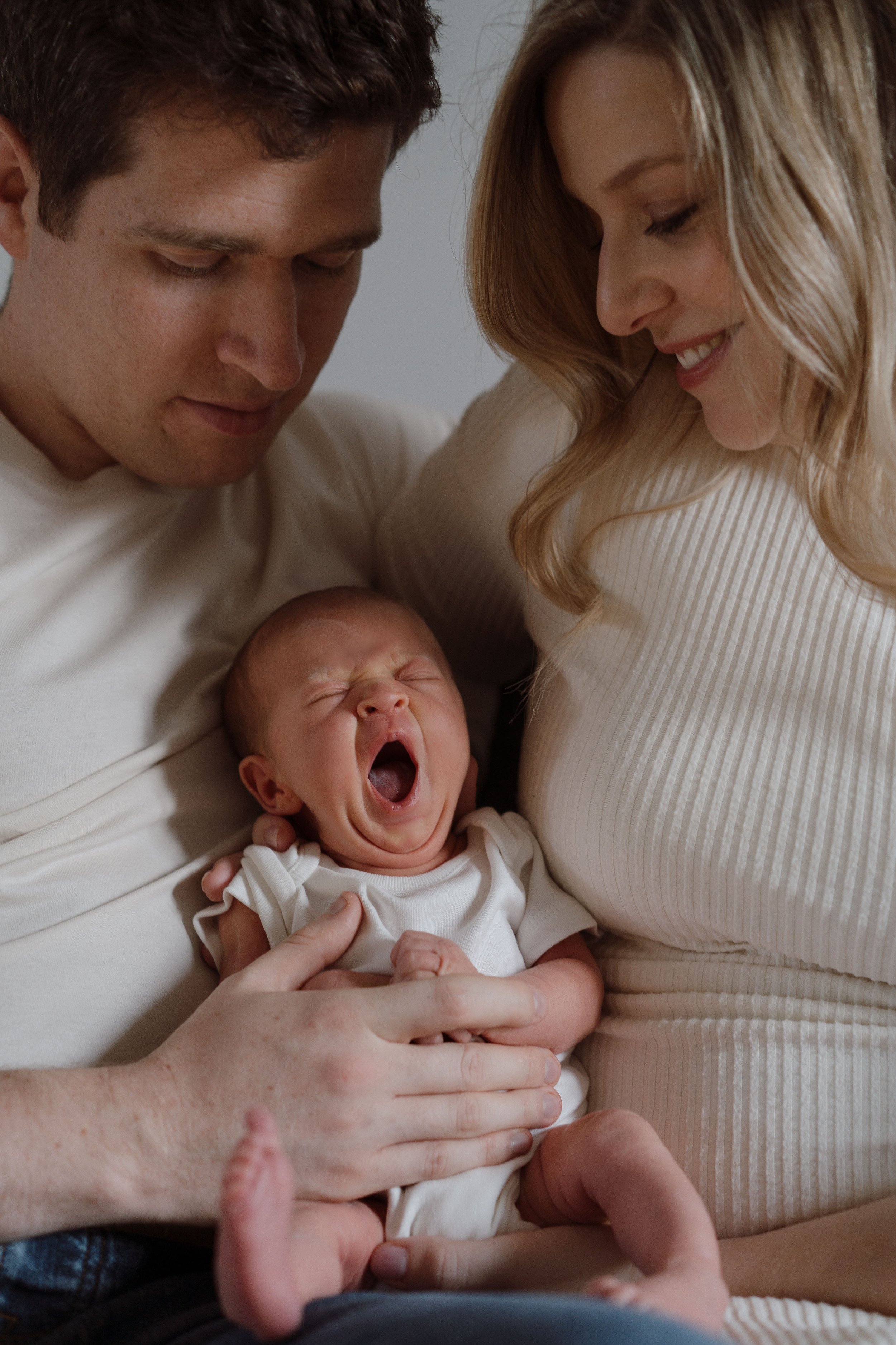 A family of three, a man, a woman, and a baby, smiling and looking at each other. The baby is yawning and being held by them.