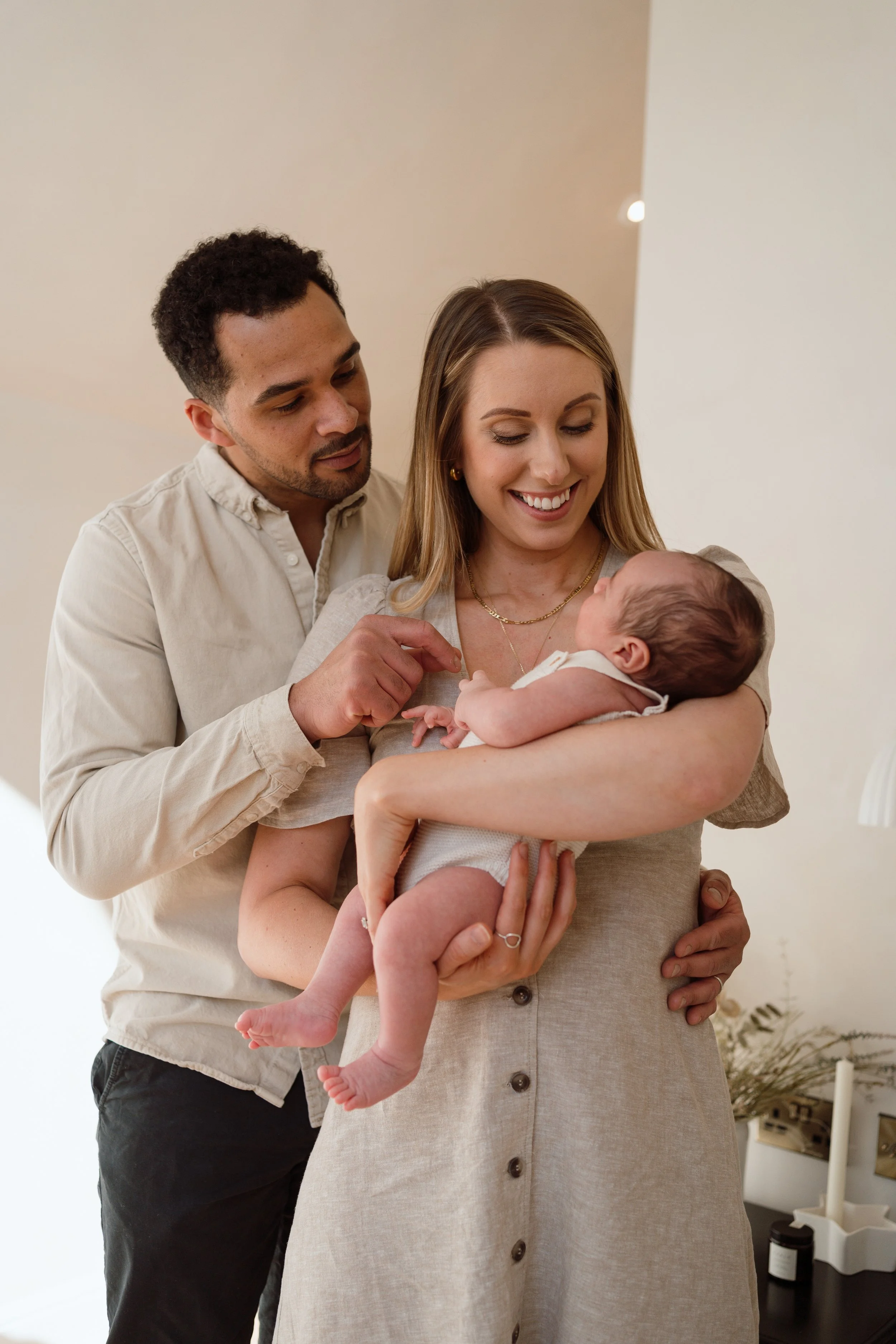 A couple holding a smiling baby girl indoors, with neutral-colored walls and some plants and candles in the background.