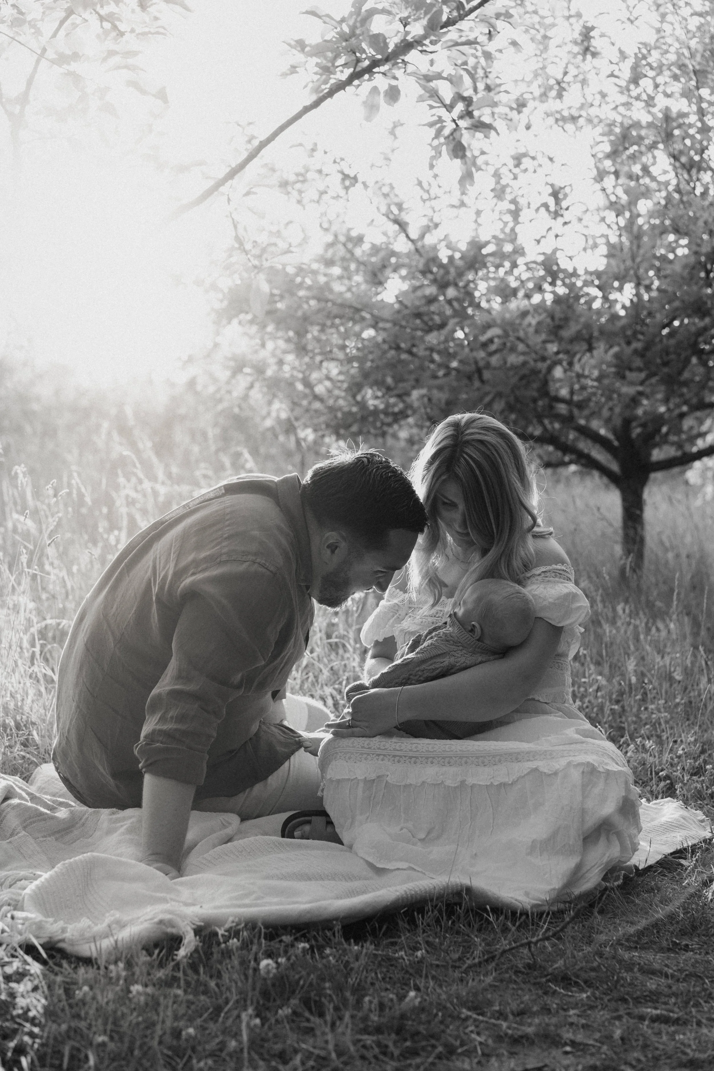 A black-and-white photograph of a family outside on a grassy area with trees in the background, showing a man, a woman, and a baby. The man is kneeling on a blanket, leaning towards the woman who is sitting and holding the baby wrapped in a blanket. 