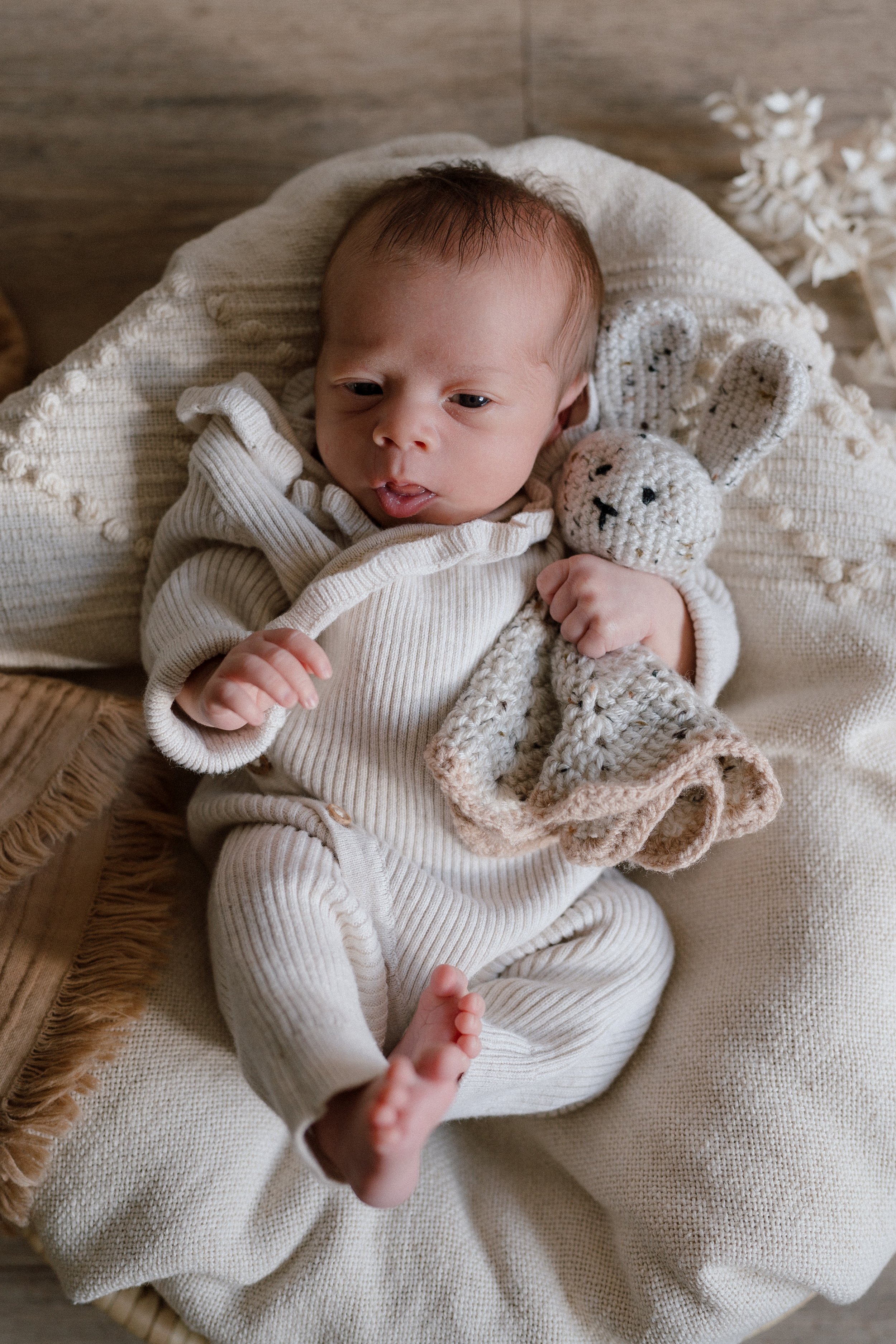 A baby lying on a cushioned surface, holding a knitted bunny toy, with a neutral-colored blanket and decor in the background.
