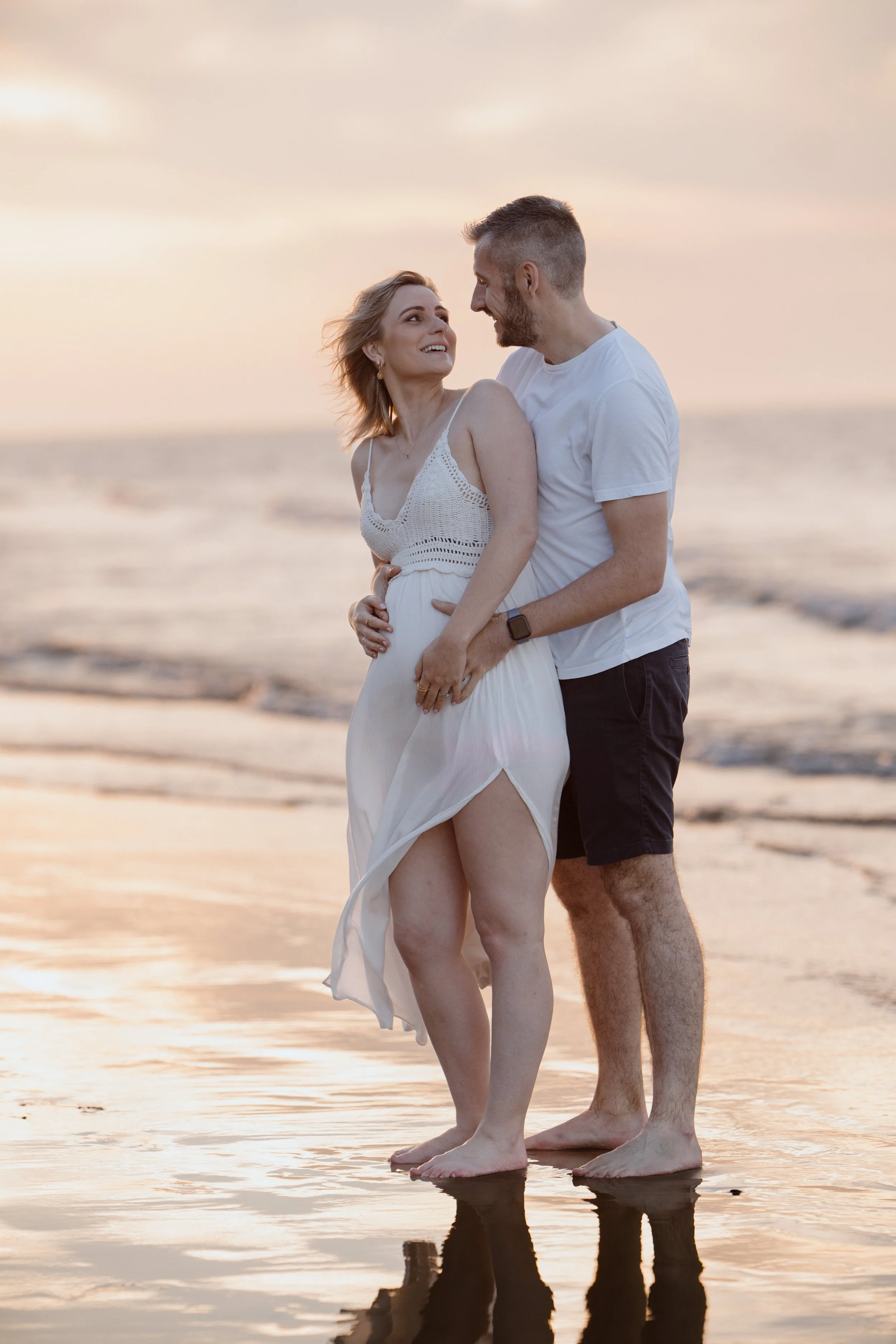 A couple standing on the beach at sunset, looking at each other affectionately, with the ocean in the background.