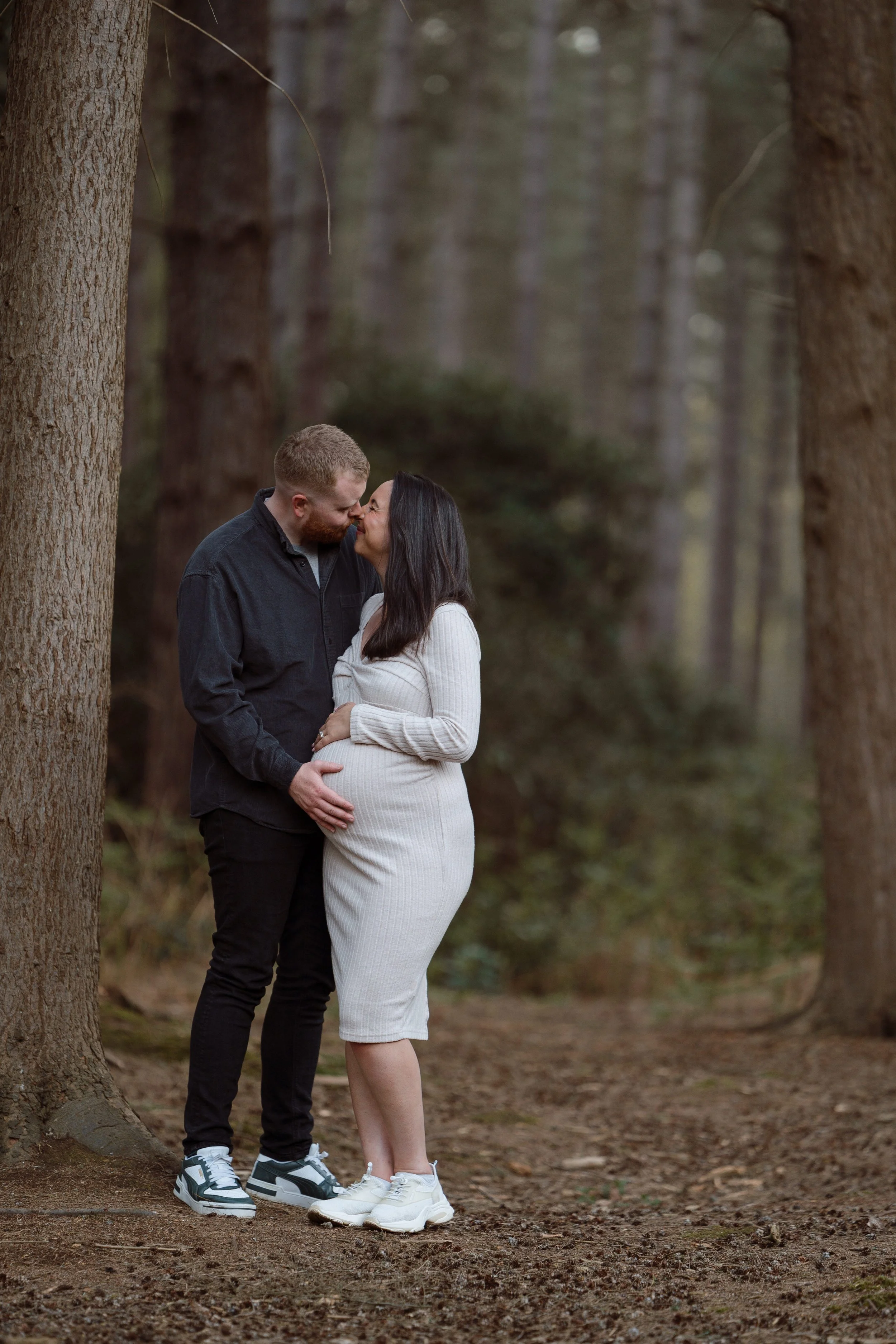 A couple in a forest standing close, touching foreheads and smiling, the woman is pregnant and holding her belly, the man gently touching her belly.